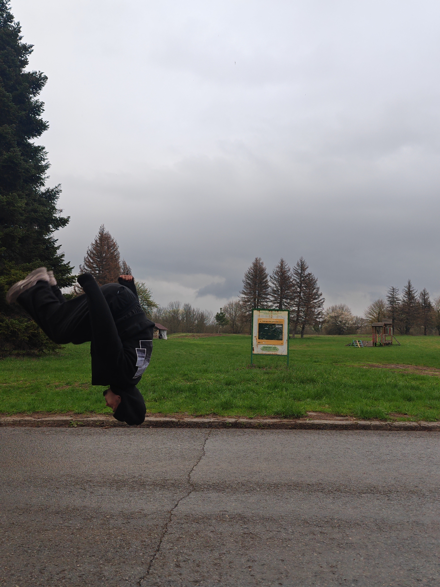 A male figure is performing a backflip on the edge of a gray asphalt road. He is dressed in black clothing and wearing gray shoes. A grassy field is behind him, marked by a large evergreen tree on the left edge. In the middle ground, there is a green and yellow map sign and a small wooden play structure. Several bare trees dot the landscape in the background. The sky is overcast with dark clouds. This location is in Shumen, Bulgaria.