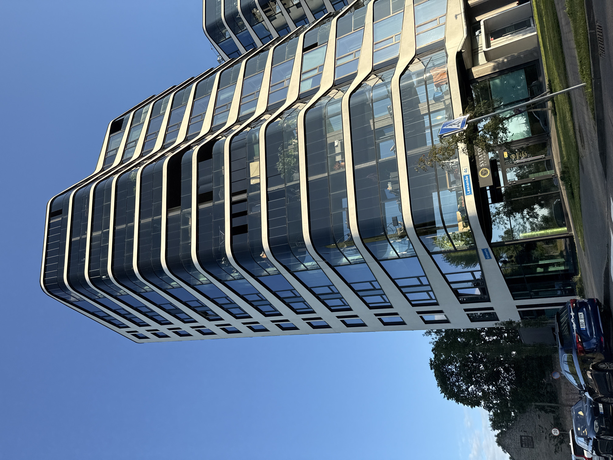 The image depicts a modern, curved building with a facade of white and dark glass, characteristic of contemporary architecture. It is situated in Tallinn, Estonia, specifically on Lasnamäe street, as indicated by a street sign visible on the building's entrance. The building has multiple stories, with each floor featuring balconies and large windows that reflect the clear blue sky and surrounding greenery.
In the foreground, several cars are parked along the street, including a dark blue SUV with license plate "680 BYK" and a black sedan. A pedestrian crossing sign and a street sign with the number "4c" are also visible. Adjacent to the modern building, an older, stone structure can be seen, suggesting a mix of historical and new development in the area. Trees and a grassy lawn border the building. The overall scene captures an urban environment under a bright, clear sky, likely during the daytime.