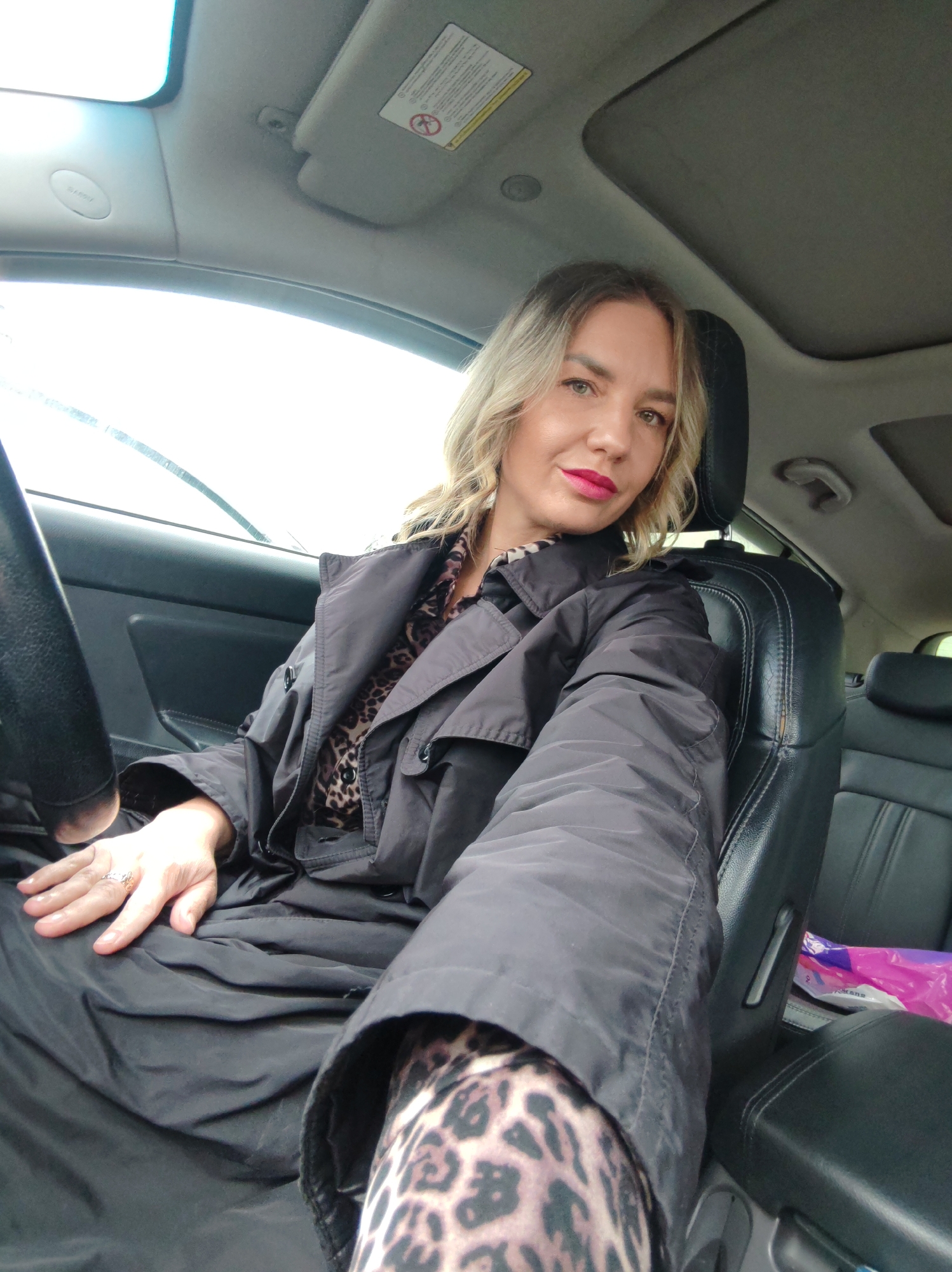 A blonde-haired woman, wearing red lipstick, is captured in a selfie while seated in the driver's seat of a car. She looks directly at the camera with a calm, composed expression. She is dressed in a dark, possibly black, trench coat or jacket, layered over a leopard print top. Her left hand, adorned with a ring, rests on her lap, where the black fabric of her coat is gathered. A leopard print legging is visible on her extended right leg.
The scene takes place inside a car, which appears to be left-hand drive, consistent with traffic laws in Russia. Bright, diffused daylight streams in from the windshield, where wipers are visible, suggesting cloudy or recently wet weather. A sunroof is also visible above. The car interior features black leather seats, a dashboard, and a steering wheel on the left.
Notable details include a sun visor on the passenger side, displaying a "WARNING" label along with an airbag safety graphic and additional text, including "AIR BAG". An "AIRBAG" indicator is also visible on the overhead console. A pink plastic bag can be seen on the passenger side floor or seat. The overall impression is of a casual moment captured in Petropavlovsk-Kamchatsky, Russia.