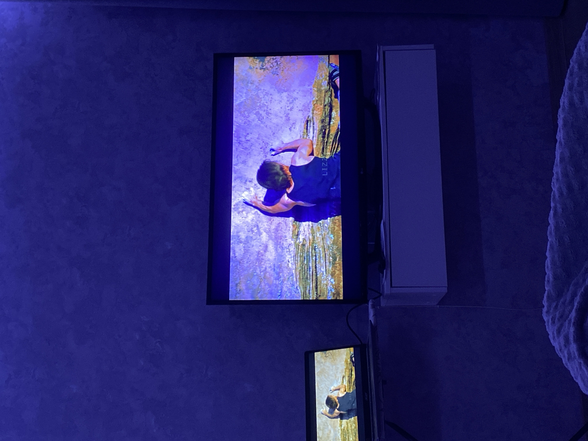 The image shows a blue-lit interior with two vertically stacked screens displaying a rock climber. The larger, upper screen shows the climber at the top of a rock face or cliff, while the smaller, lower screen offers a closer view of the climber's upper body. A white cabinet is adjacent to the upper screen. The location of the image is Зубово, Russia.