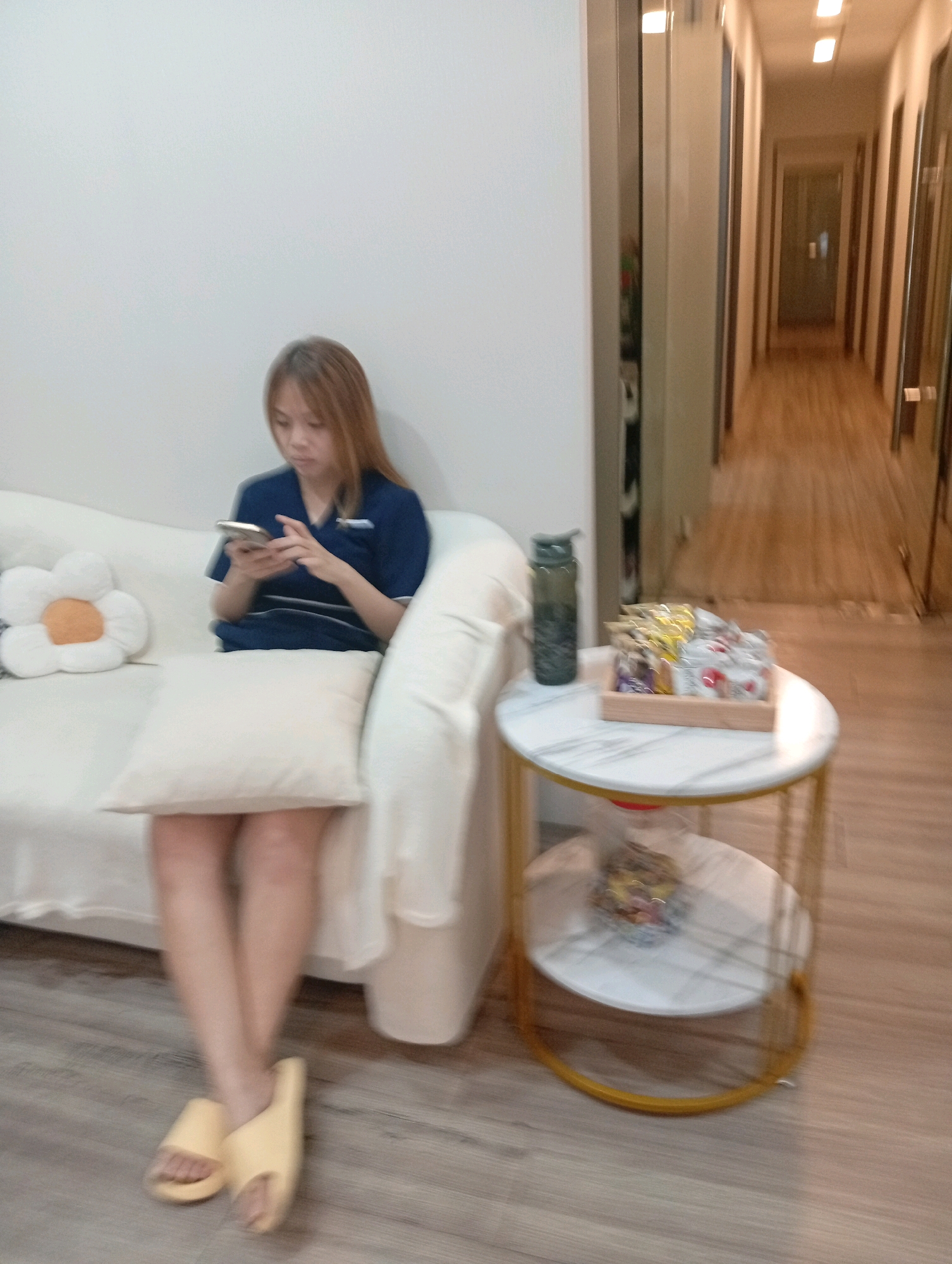 A young Asian woman with shoulder-length, light brown hair is seated on a white couch, looking down at her phone. She is wearing a dark blue v-neck top with a white emblem on the left chest and dark blue bottoms. A white throw pillow with a flower-shaped cushion featuring an orange center is to her left. Another white throw pillow is placed in front of her. The couch is positioned against a white wall. To the right of the couch, there is a two-tiered side table with a marble-patterned top and a gold frame. On the top shelf of the table sits a dark green water bottle and a wooden tray filled with several individually wrapped snack items. The bottom shelf has a similar marble-patterned top. The scene appears to be indoors, possibly in a waiting area or lounge. In the background, a long hallway with multiple doors is visible, suggesting a commercial or institutional setting. The floor is a light-colored wood. The overall impression is one of a clean, modern interior.