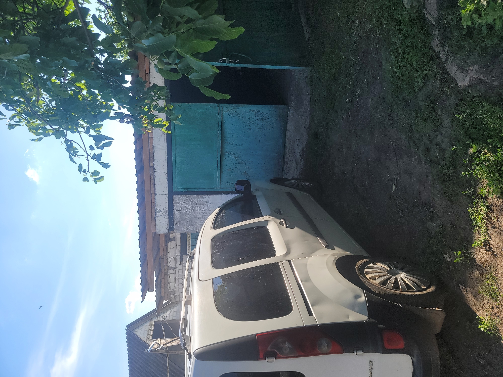 In Vovkivka, Ukraine, a light-colored panel van is parked on a dirt and grass area in front of a detached garage. The garage has a weathered teal-colored double door that is partially open, revealing a dark interior. The van is positioned to the left of the garage, with its rear facing towards the viewer and its side visible. Overhanging the scene from the right is a large tree with lush green leaves, casting shadows on the ground. The sky above is a clear blue with a few wispy white clouds. The overall environment suggests a rural or semi-rural setting, likely a private property.