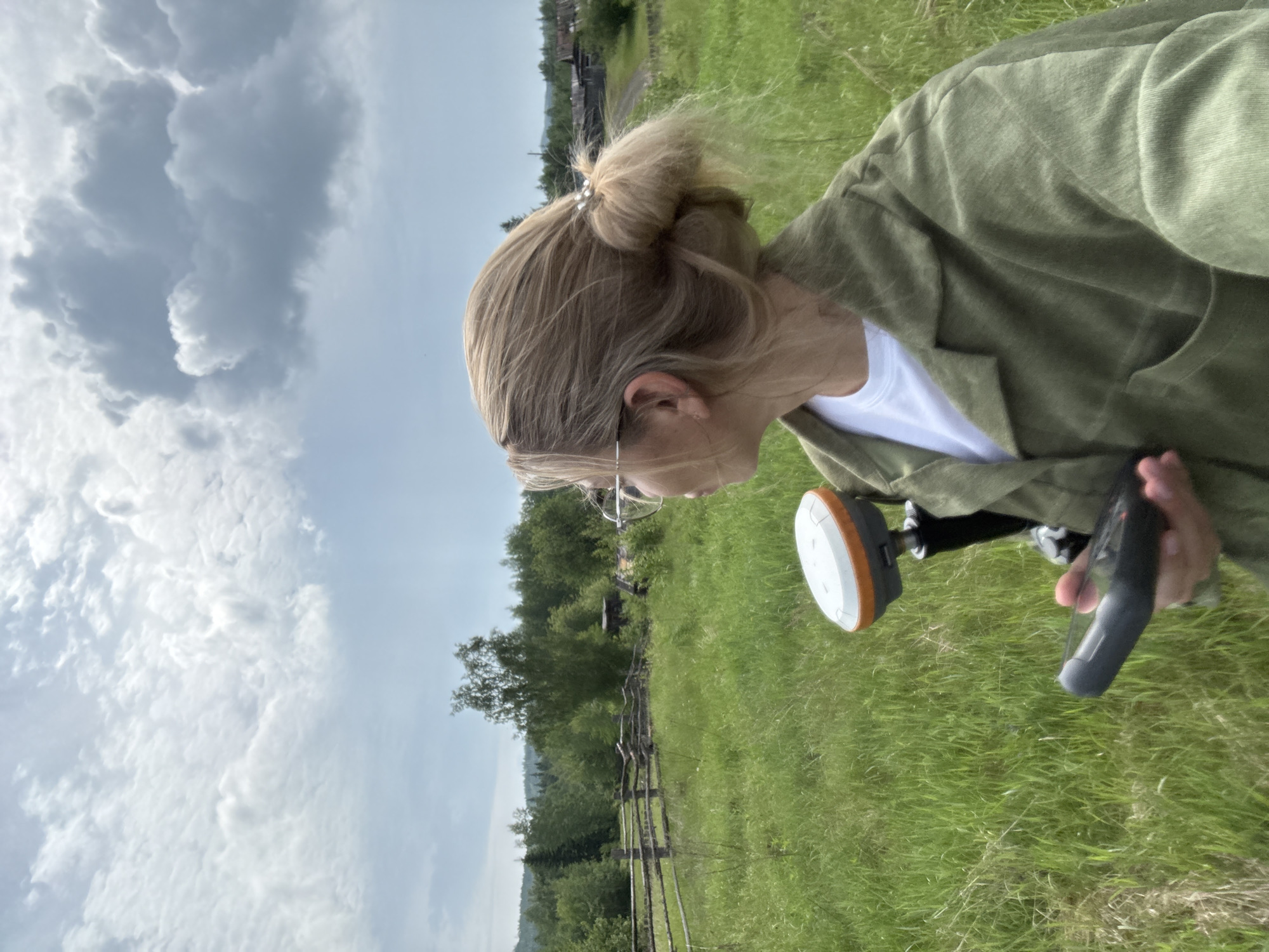 A woman with blonde hair tied in a bun is standing in a grassy field, holding a surveying device. She is wearing glasses and a green jacket over a white t-shirt. The background shows a wooden fence, trees, and a few rustic buildings. The sky is cloudy. The location is identified as Badarminsk, Russia.