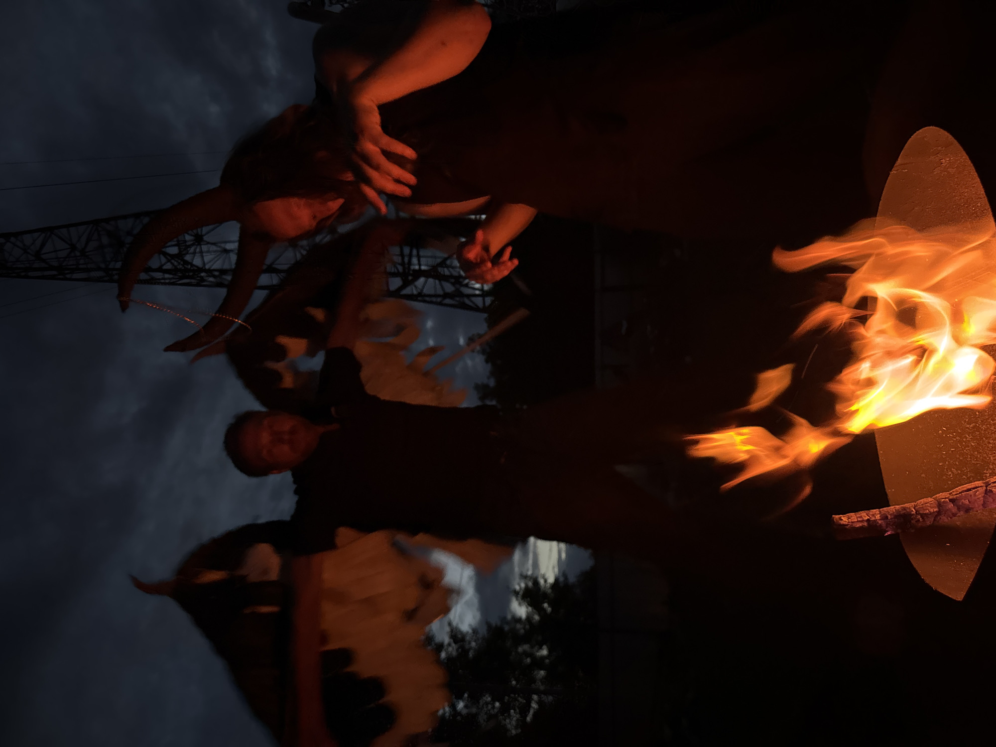At dusk in an outdoor area, likely on the outskirts of Chaikovsky, Russia, two individuals are engaged in an activity near a bonfire. The scene is dimly lit, with the sky appearing dark and overcast.
The individual on the left is wearing what appears to be large, feathery wings. They are dressed in dark clothing and have their arms outstretched. Their face is partially obscured.
The individual on the right is wearing a headdress with prominent horns. They are also dressed in dark clothing. Their hands are visible, and they appear to be holding or interacting with a thin chain or necklace. Their face is also shadowed.
In the foreground, a fire burns in a round metal receptacle. The flames are bright orange and cast a warm glow on the immediate surroundings. Behind the individuals, a tall, lattice-like structure, possibly a power pylon, is visible against the dark sky. Trees are silhouetted in the background, suggesting a natural or semi-rural setting within the city limits. The overall atmosphere is dramatic and somewhat ominous.