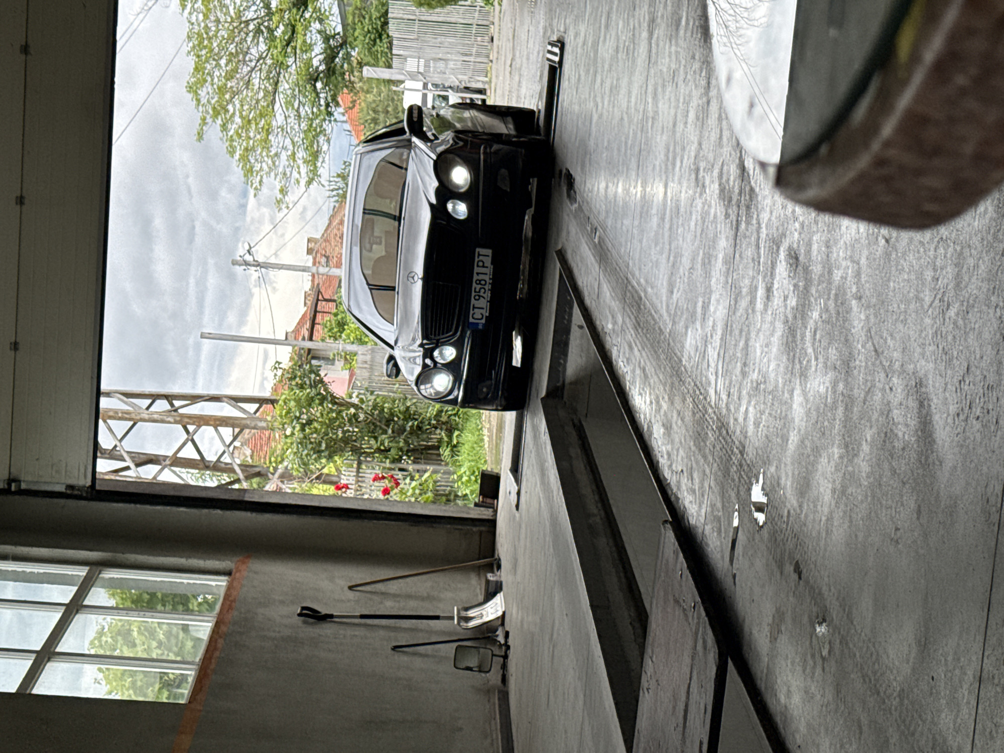 The image depicts a black Mercedes-Benz parked inside a garage or workshop. The car has a license plate reading "CT 9581 PT." Outside the garage door, there's a glimpse of greenery, residential buildings, and utility poles under a cloudy sky. Inside the garage, there are cleaning tools like a broom and a squeegee against the wall, as well as a mirror. The floor is concrete with visible tire tracks. The image location cannot be pinpointed to Stara Zagora, Bulgaria, without additional information or context.