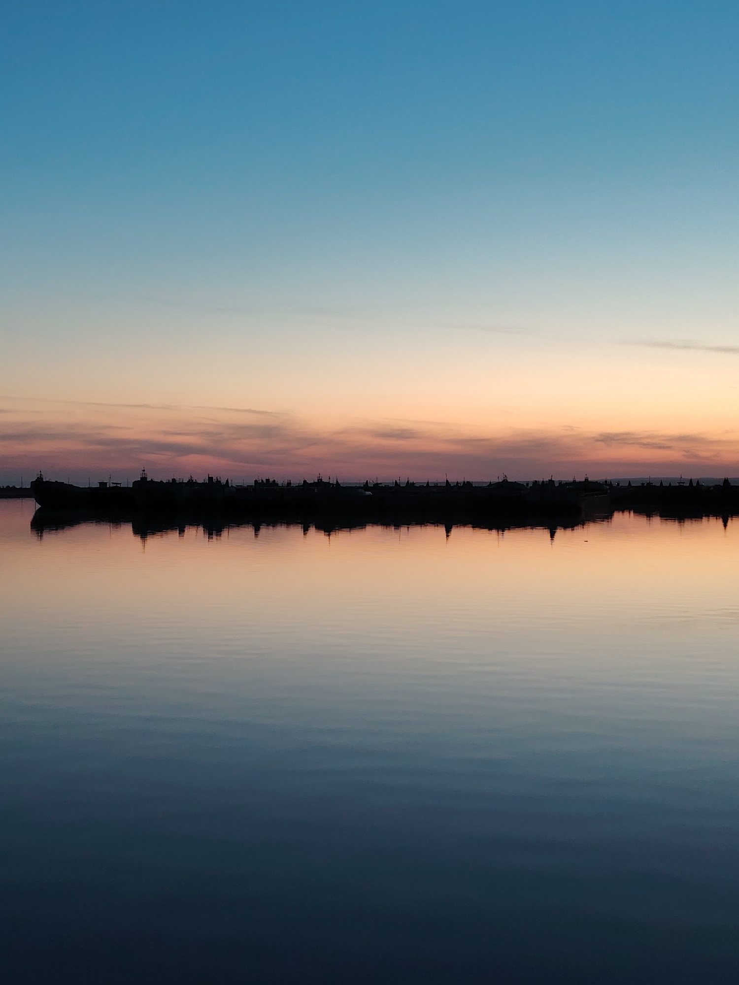 The image depicts a tranquil body of water at dusk. The sky transitions from blue to orange and pink hues near the horizon. A silhouetted landmass or structure, possibly a collection of ships, lies along the horizon line. The water surface is smooth, reflecting the colors of the sky and the silhouetted objects, creating a symmetrical effect. The location is likely in Chaikovsky, Russia. There are no visible people in the image.