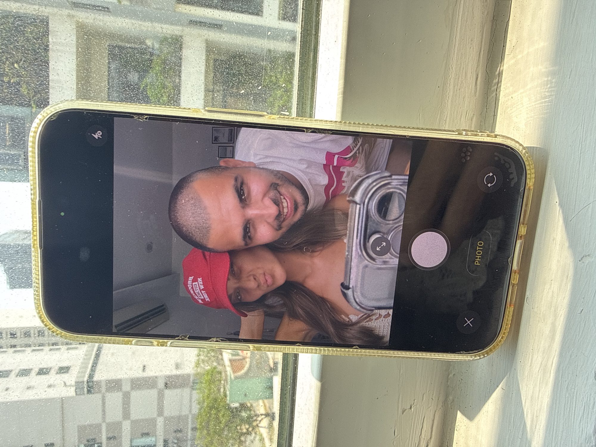 A smartphone displaying a selfie of a man and a woman is resting on a windowsill. The man has dark hair, a short beard, and is wearing a white t-shirt with a pink design. The woman is wearing a red baseball cap with white writing and a light-colored top. The selfie is taken indoors, likely inside an apartment or hotel room.
Outside the window, there is a view of Đà Nẵng, Vietnam, including high-rise buildings and greenery. The exterior of the building has windows and a light-colored facade. The light is bright and sunny, casting shadows on the windowsill.