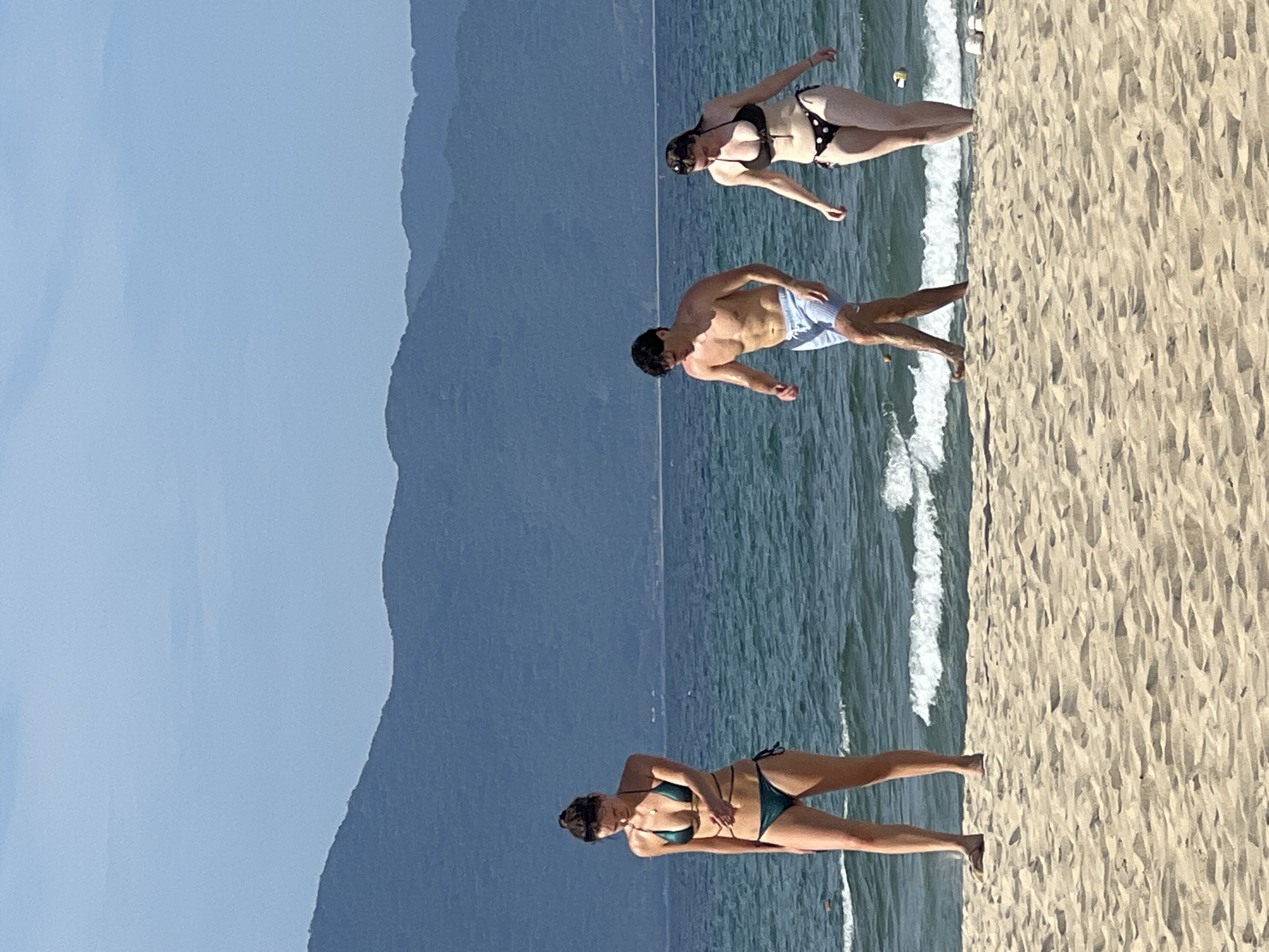 The image captures a scenic beach in Đà Nẵng, Vietnam, with the majestic Sơn Trà Peninsula mountains forming a picturesque backdrop. Three individuals are seen enjoying the sandy shore and the turquoise waters. On the left, a woman in a teal bikini stands facing away from the camera, her gaze directed downwards. To her right, a man in light blue swim trunks walks towards the right, his torso bare and tanned. Further to the right, a woman in a black and white polka dot bikini walks out of the water, her head tilted down as she steps onto the sand. The ocean exhibits gentle waves, and the clear blue sky suggests a pleasant day. The sand is a light golden hue, marked with subtle footprints and ripples.