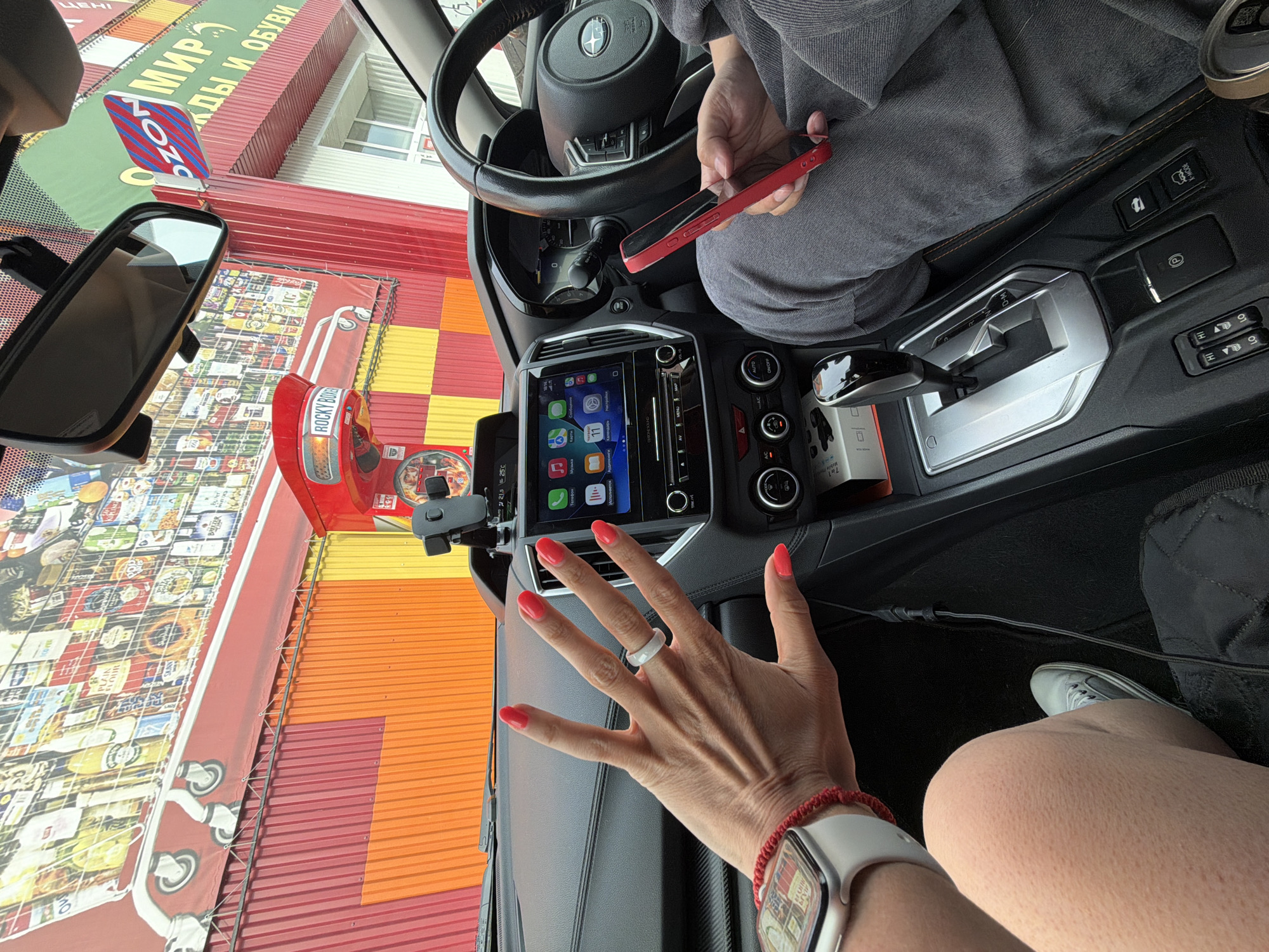 The image shows the interior of a car, likely a Subaru, with a person's hand and leg visible. The hand, adorned with bright red nail polish and a white ring, is raised, palm facing the camera, revealing a smartwatch on the wrist. The person's leg, clad in grey trousers, is bent, and they are holding a red smartphone. The car's dashboard features a prominent touchscreen display showing a car interface with various app icons and a temperature reading of 25°C. Outside the car, a colorful building facade is visible, featuring a large graphic of a shopping cart filled with products, and signage that includes the word "OZON" and Russian text that translates to "world of goods and shoes." The presence of "OZON" and the architecture suggest a location in Russia. Given that this is a known retail chain in Russia, and considering other details that may have been present in a broader context of this image, the scene is likely at an OZON pickup point or store in Birobidzhan, Russia.