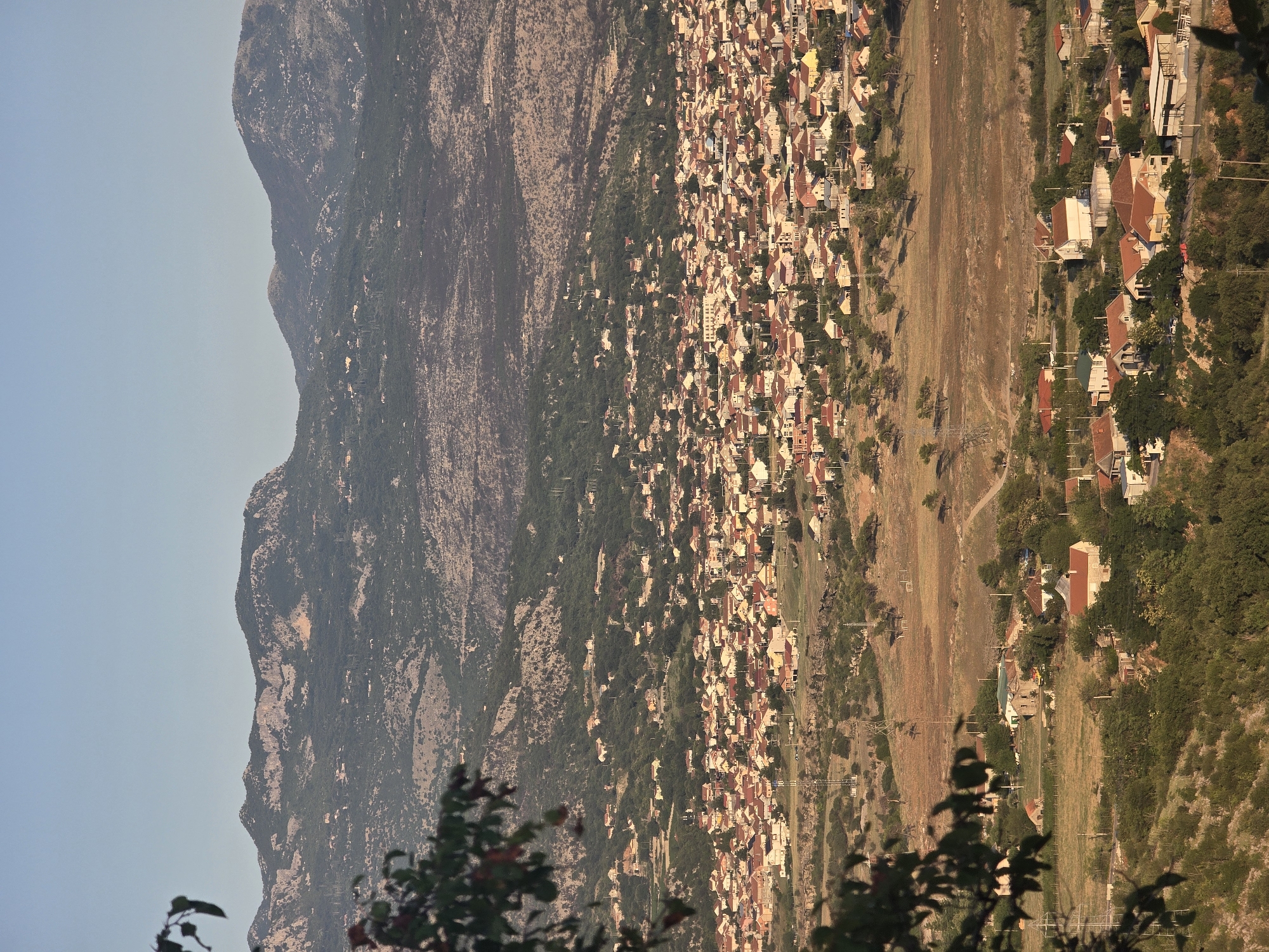 An aerial, vertical, medium shot captures a panoramic view of a densely populated town nestled in a valley below rolling hills and rugged, forested mountains under a clear, pale blue sky. The town, characterized by a uniform architectural style of low-rise houses with terracotta-tiled roofs, dominates the middle ground. The houses are primarily light-colored, with some exhibiting pastel hues, and are tightly clustered together, interspersed with trees and small open spaces. Power lines and electrical poles are visible throughout the town and the surrounding landscape.
In the foreground, the scene is partially obscured by the dark leaves and branches of a tree, suggesting the vantage point is from a higher elevation. The terrain transitions from the built-up area to drier, less vegetated hills dotted with sparse trees and rocks. Further in the distance, the imposing mountains form a dramatic backdrop, their textured surfaces a mix of rocky outcrops and dark green forests. The overall lighting suggests it is either early morning or late afternoon, casting soft shadows across the landscape.