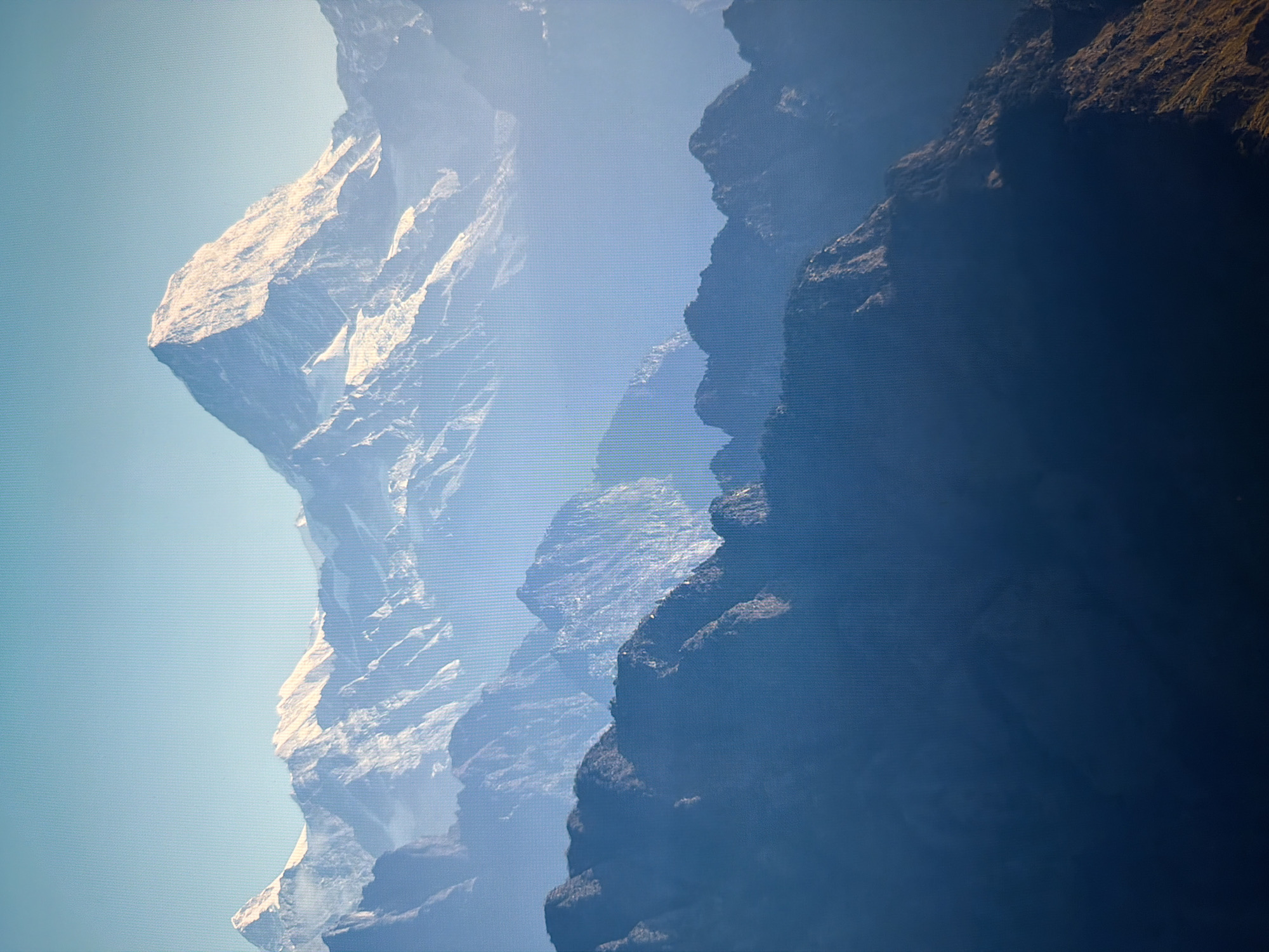 This is an image of a majestic mountain range. The foreground features dark, silhouetted mountain peaks, gradually giving way to lighter, mist-shrouded slopes in the mid-ground. The background is dominated by a towering, snow-capped mountain peak, brightly illuminated by sunlight, with clear blue sky above. The scene conveys a sense of scale and natural grandeur. There are no elements in this image that suggest a location in Kyiv, Ukraine.