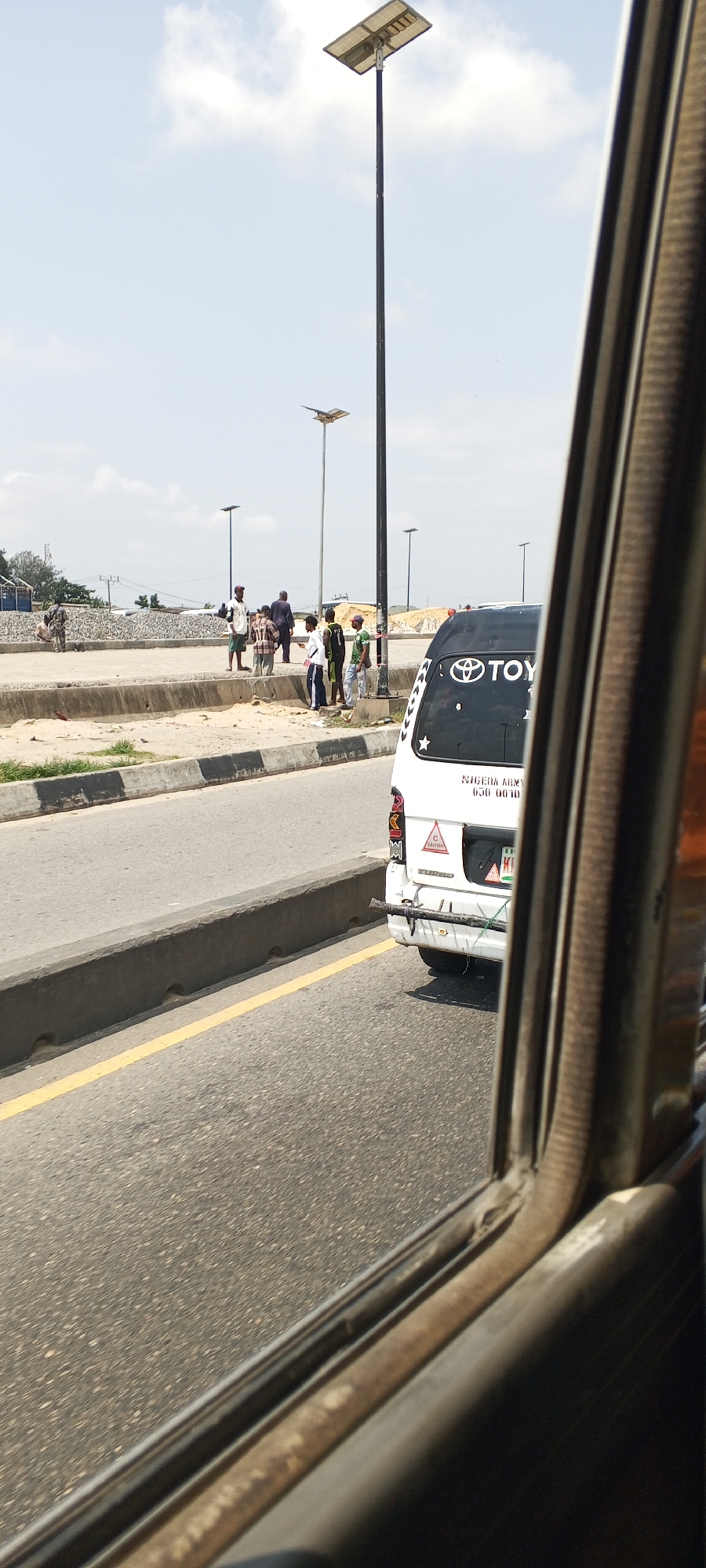 The image shows a street scene in Unknown, Unknown on a sunny day. A white Toyota van with "TOYOTA" logo on the back is parked on the right side of the road. The van has "NIGERIA ARMY" and a license plate number "630 0010" written on its rear. Several people are standing on the side of the road near some construction materials, possibly waiting for a ride or observing the surroundings. There are solar-powered streetlights visible in the background. The photo is taken from inside a vehicle, looking out through the side window.