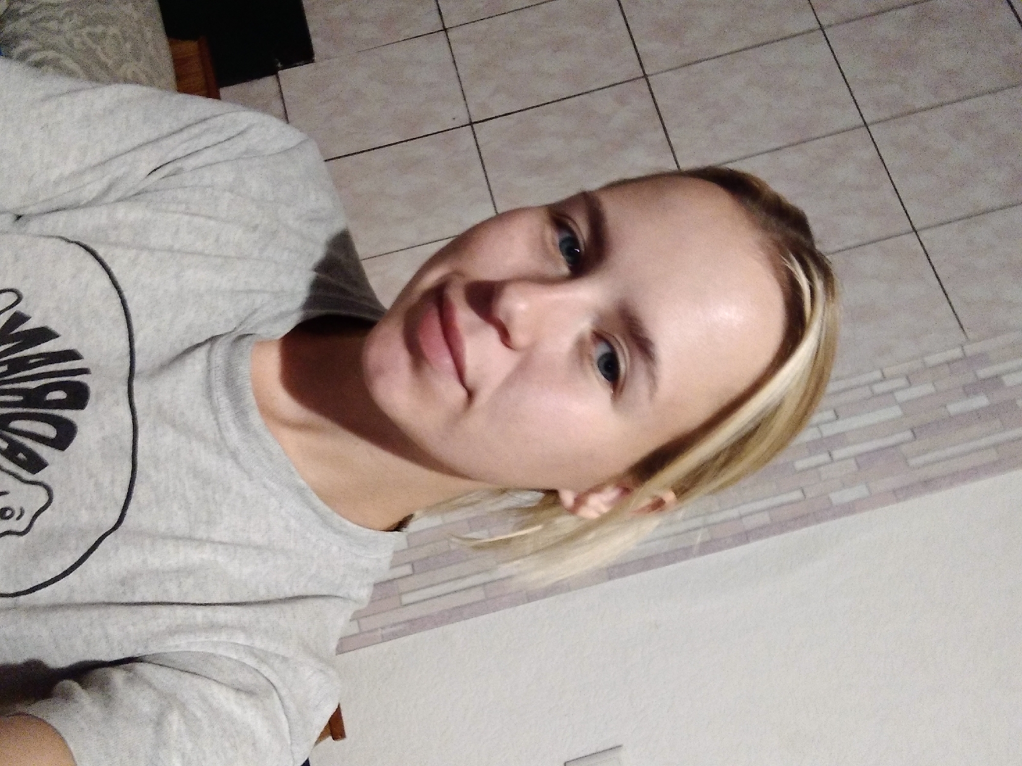 A young woman with blonde hair and blue eyes is taking a selfie indoors, with a tiled wall and a patterned wall visible in the background. She is wearing a grey t-shirt with a circular graphic design. The environment suggests a residential setting, possibly in the city of Radisne, Ukraine, given the location information provided, although the image itself does not contain any specific identifying markers of that location. The lighting is dim, casting a slight shadow on her face.