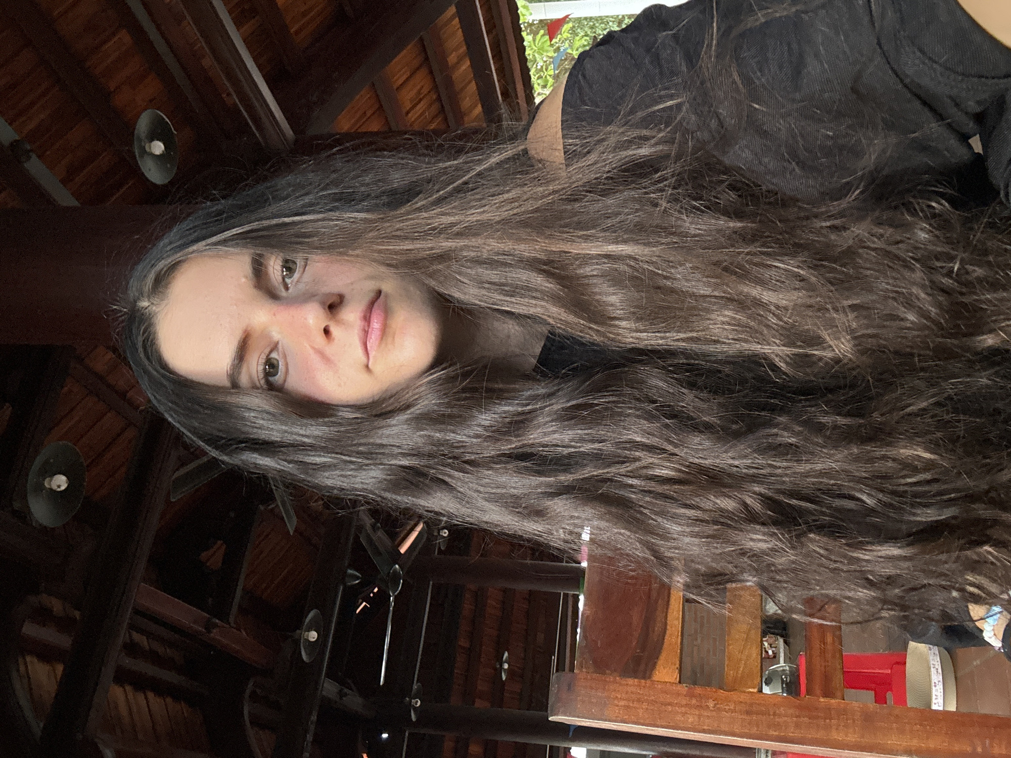 A woman with long, wavy brown hair and light skin is looking at the camera. She is wearing a black shirt. The background consists of a dark, wooden structure with visible ceiling beams and what appears to be lighting fixtures. In the distance, hints of greenery and a red object are visible through openings in the structure. The location appears to be indoors within a wooden building structure. The building looks to be located in Mũi Né, Vietnam.