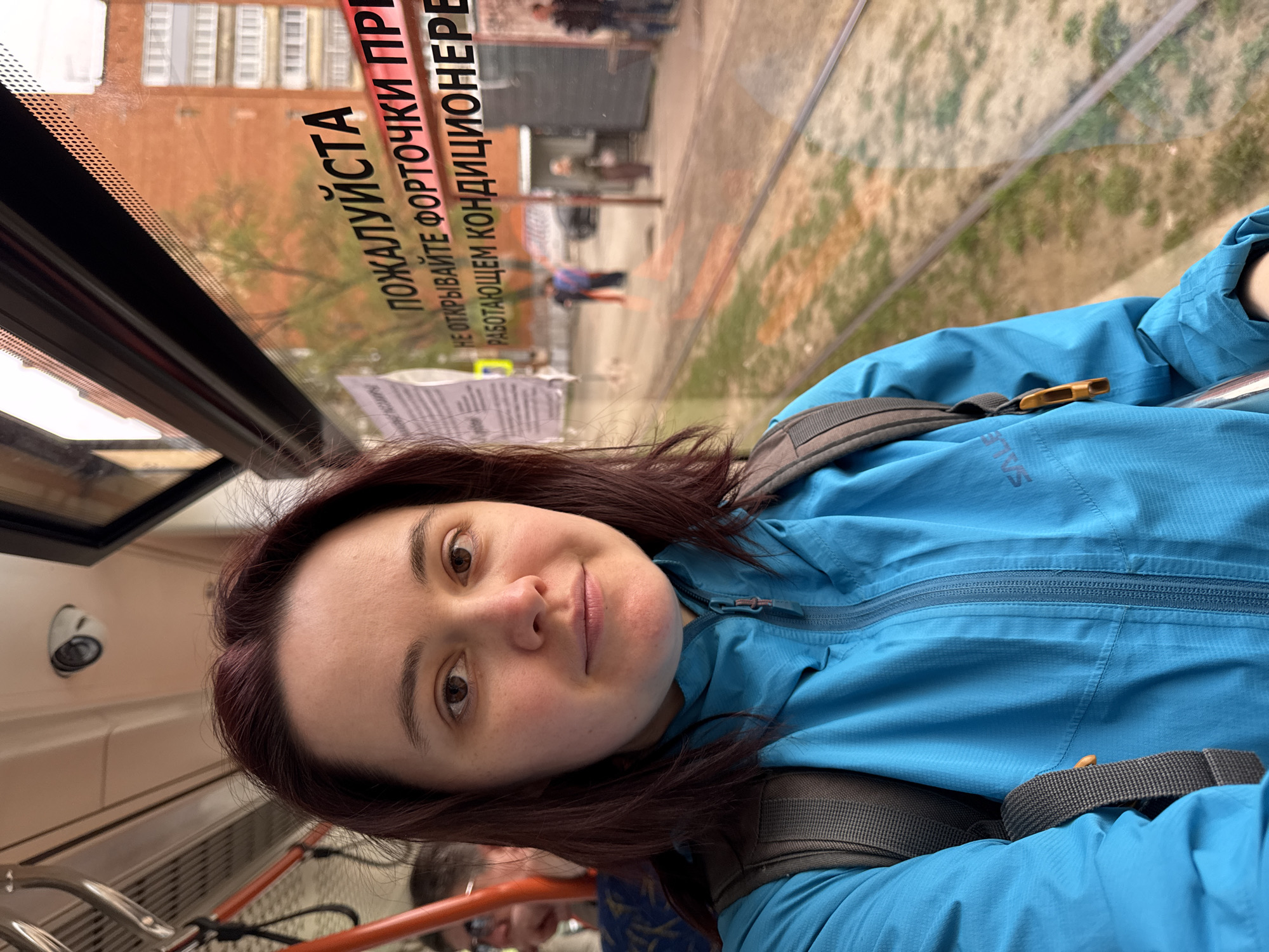 A woman with reddish-brown hair is taking a selfie inside a tram or similar public transport. She is wearing a blue jacket and backpack. Outside the window, a street scene is visible, including a building, trees, and pedestrians. A sign on the window reads "ПОЖАЛУЙСТА НЕ ОТКРЫВАЙТЕ ФОРТОЧКИ ПРИ РАБОТАЮЩЕМ КОНДИЦИОНЕРЕ," indicating that the tram has air conditioning and passengers are requested not to open the windows. The location is potentially Izhevsk, Russia. Another person is seen in the background.