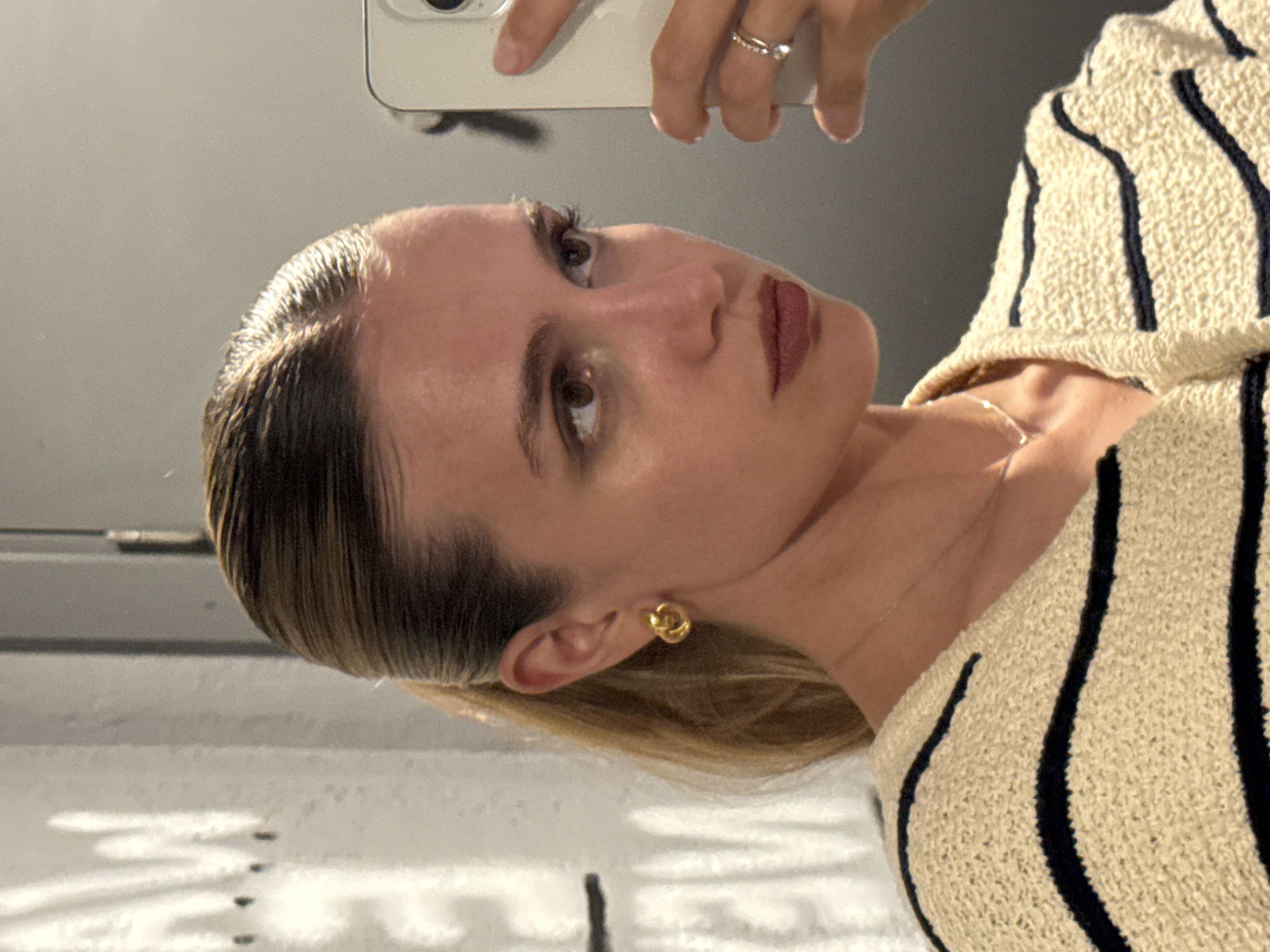 This mirror selfie captures a young adult female from the chest up, with her head slightly tilted, looking upwards and to her right. Her hair is styled in a sleek, pulled-back, wet-look fashion, showcasing a transition from light brown roots to blonde ends. She has light-colored eyes, well-defined eyebrows, and noticeable eyelashes, with her lips painted in a dark reddish-brown shade. Her skin appears smooth with a natural glow.
She is dressed in a textured, light beige or cream-colored knit sweater featuring prominent dark vertical stripes. A delicate gold-toned chain necklace is visible around her neck, and she wears a small, gold knot-style earring in her left ear. A silver-colored ring with a small clear stone adorns her left ring finger. The upper part of a light-colored smartphone, which she is holding to take the picture, is visible in the reflection above her head.
The setting is an indoor space, likely a bathroom or dressing area, as suggested by the plain light grey background and a white, subtly textured surface with faint patterns reflected in the lower part of the mirror. The subject's expression is calm and contemplative. There is no visible text in the image. The location context for the image is Tbilisi, Georgia.