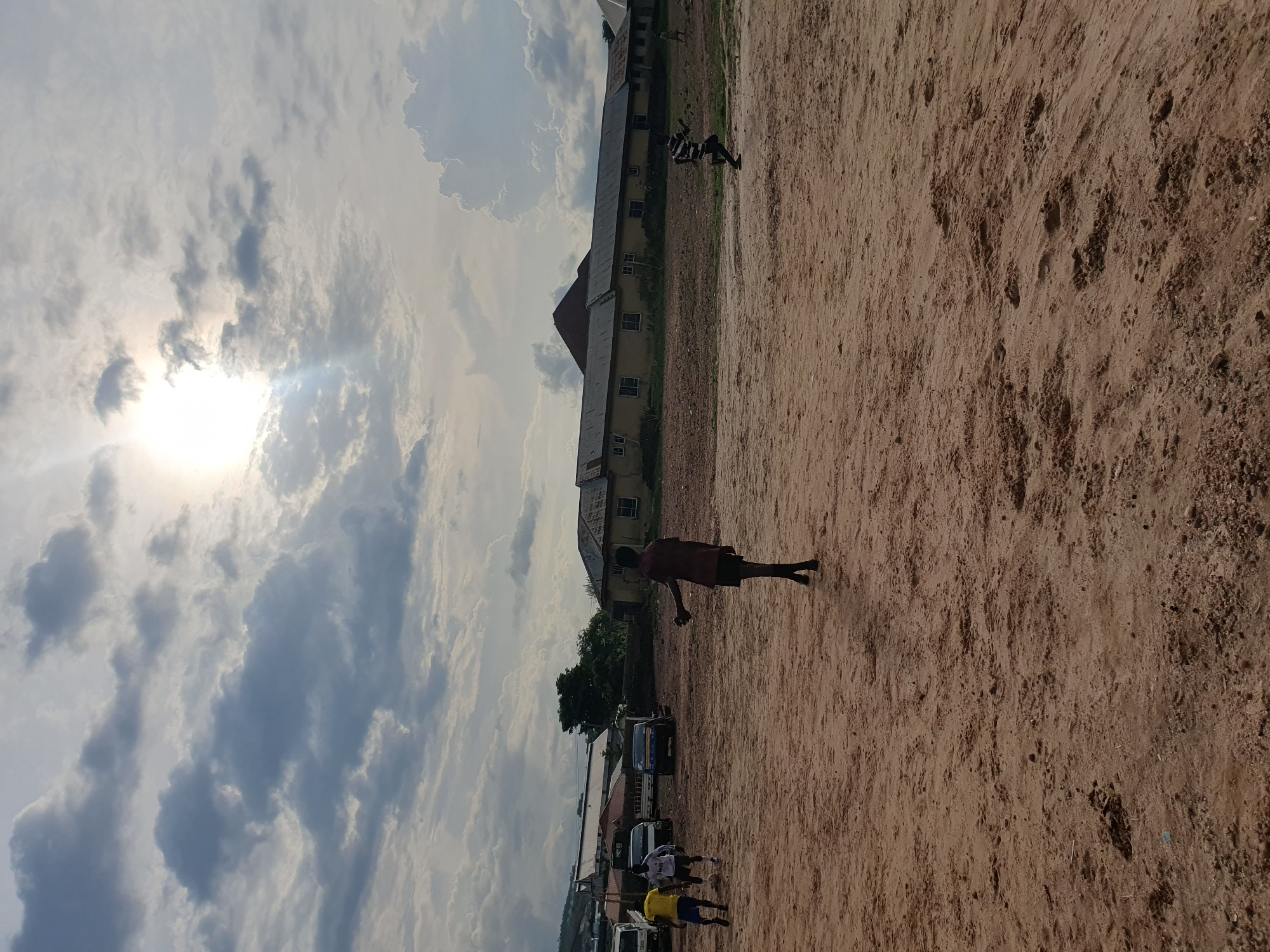 The image depicts a field in Lokoja, Nigeria, under a partly cloudy sky with the sun visible. The ground is dry and sandy. A group of people, including children, are present, some appearing to be playing. A few vehicles are parked alongside a building. One individual is prominently positioned in the foreground, in motion. There is a building behind them.
