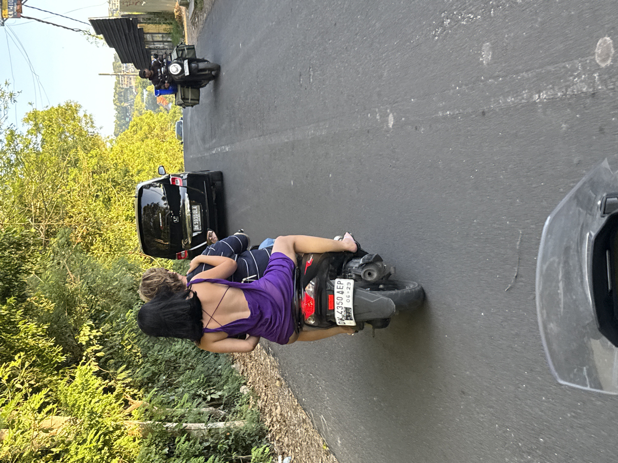 The image depicts a road scene in Kuta Selatan, Indonesia. A woman in a purple dress is riding as a passenger on a scooter, wearing no helmet or shoes, and hugging the driver, who is wearing striped pants. The scooter's license plate reads "DK 4350 AEP 06.29". A black car with the plate "DK1030GLA" is nearby, and a motorcycle is visible ahead on the road. Green foliage lines the side of the road, and a partial view of a helmet camera is in the foreground.