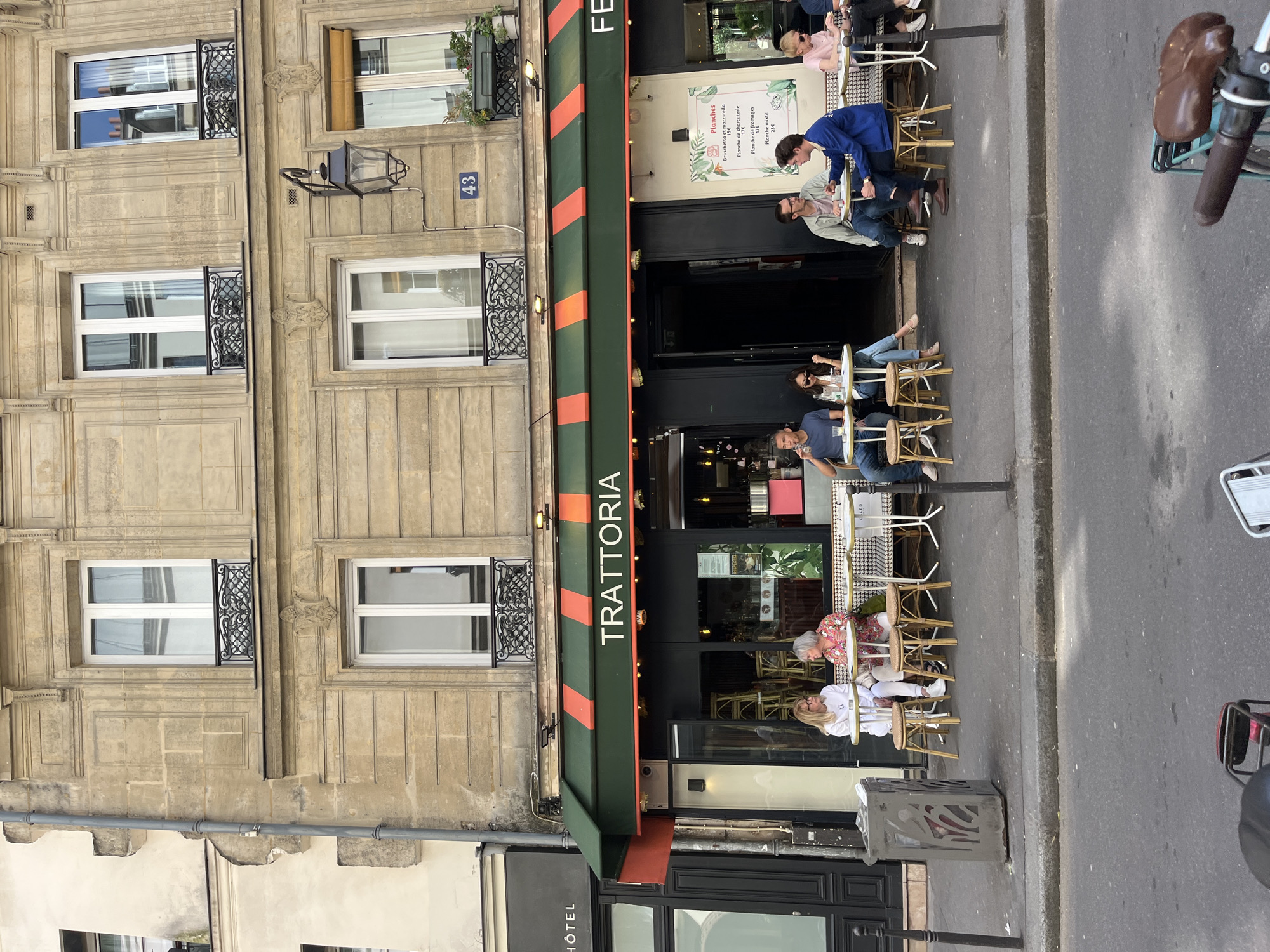 The image captures an outdoor scene in Paris, France, showcasing a restaurant named "Trattoria" with an awning in dark green and orange. Several people are seated at tables on the sidewalk, engaged in dining and conversation. The building facade is of a traditional Parisian design, featuring stone cladding, windows with wrought-iron details, and a building number "43". A menu board outside lists various "Planches" (platters). Part of a bicycle and a street trash receptacle are also visible on the right side of the frame, suggesting an urban setting. Additionally, a sign for a "Hôtel" is visible adjacent to the Trattoria.