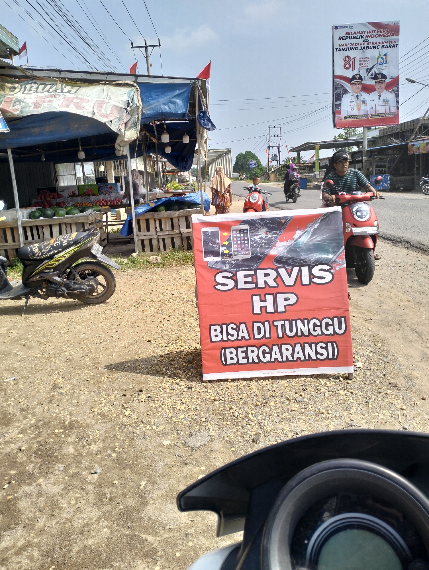 A daytime outdoor shot captures a street scene in Pematang Lumut, Indonesia. In the foreground, a red and white sign advertises "SERVIS HP BISA DI TUNGGU (BERGARANSI)" with images of cracked phone screens. A portion of a motorcycle's dashboard is visible at the bottom. To the left, a parked black motorcycle is in front of a fruit stand. The fruit stand, covered by a blue tarp and featuring a banner with the word "BUAH," displays various fruits, including watermelons and apples. Beyond the fruit stand, several people are present, some walking and others riding red scooters. In the background, a large billboard celebrates "SELAMAT HUT KE-81 REPUBLIK INDONESIA" and "HARI JADI KE-61 KABUPATEN TANJUNG JABUNG BARAT" with images of local officials. Overhead, power lines crisscross the sky under a clear, blue sky. The ground is covered with gravel and dirt.