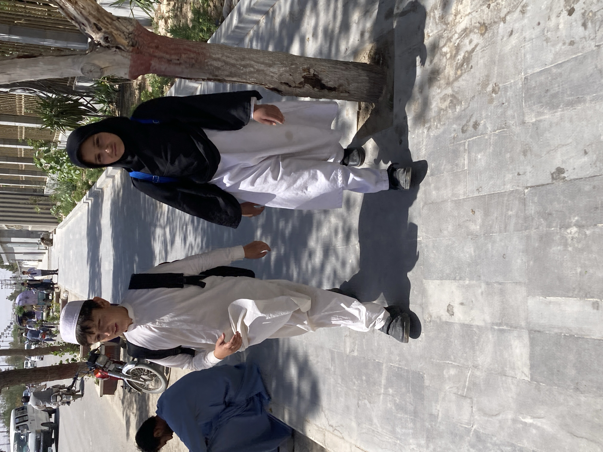 The image, rotated 90 degrees counter-clockwise, captures an outdoor scene in what appears to be an urban or semi-urban setting in Quetta City Tehsil, Pakistan, during daylight hours. The strong shadows suggest a sunny day, likely mid-morning or afternoon.
The primary subjects are two young individuals standing on a paved sidewalk. On the left, a girl wears a black head covering (hijab), a black top, and white pants, with a blue strap visible on her shoulder, possibly from a backpack. She looks directly at the camera. To her right, a young boy is dressed in a white traditional shalwar kameez-style outfit and a white cap, also looking towards the camera with a slight smile. He has a black strap across his shoulder.
Behind the girl and boy is a tree with a prominent trunk, some exposed roots, and a patch of red paint on its lower section. The sidewalk is made of light-colored, possibly concrete or stone, rectangular pavers, showing some wear and stains.
In the lower right foreground, a third person is partially visible, bent over and wearing a blue traditional outfit. The background reveals a street scene with other pedestrians, parked vehicles including a motorcycle, and distant buildings, consistent with a busy street environment. No visible text is present in the image.