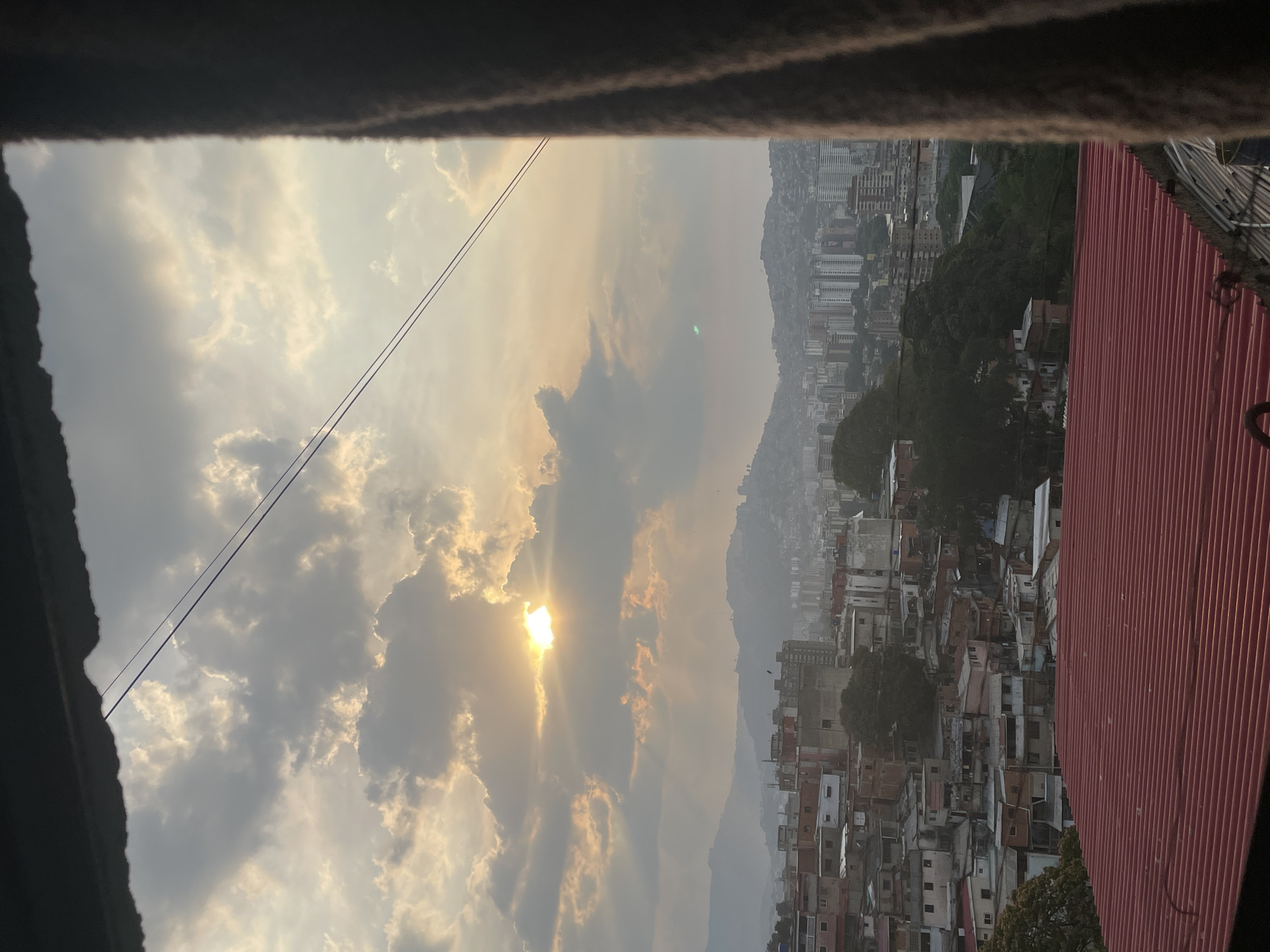 The image depicts a hazy sunset over a densely populated urban area, likely a hillside neighborhood in Caracas, Venezuela. The view is framed by a window or opening. In the distance, tall buildings can be seen rising from the terrain. In the foreground, crowded buildings are visible on a slope. The sky is filled with clouds, and the sun is partially obscured, casting rays through the clouds. High voltage wires are crossing the sky. A corrugated roof is visible.