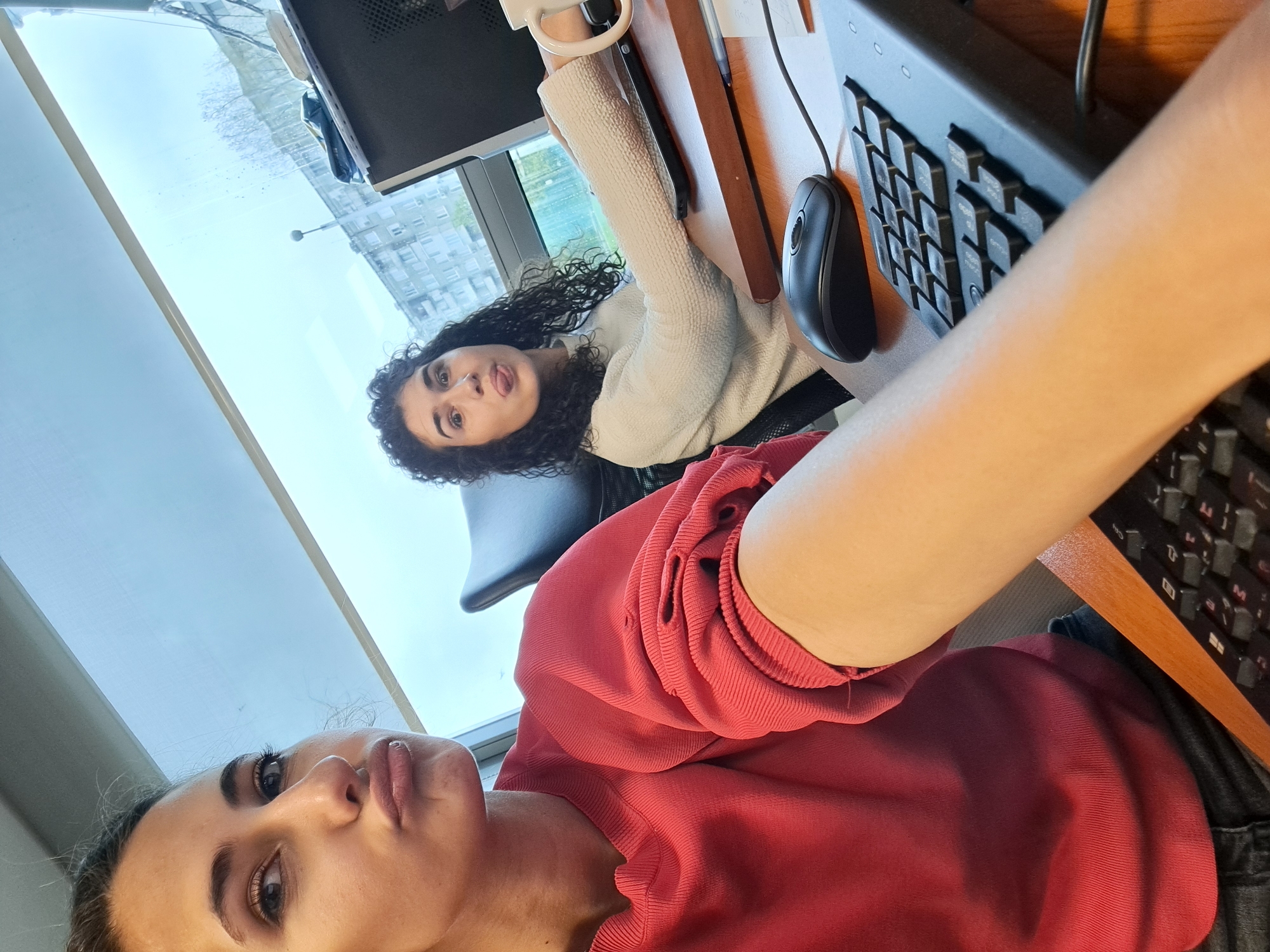 The image is a low-angle, rotated selfie taken indoors, featuring two young women in what appears to be an office or study environment.
In the foreground, a woman with dark hair, wearing a red long-sleeved top, is taking the photo. She looks directly into the camera with her lips slightly pursed. Her right arm extends into the shot, holding the camera.
Behind her, seated at a wooden desk, is another woman with dark, curly hair, wearing a textured white long-sleeved top. She is looking towards the camera with a playful expression, her tongue slightly out, and holds a white mug in her right hand.
On the desk, parts of a black computer monitor, a black keyboard, and a black computer mouse are visible. The woman in the background is seated in a black office chair.
Behind the desk and the women, a large window reveals an outdoor scene. Through the window, multi-story buildings and some faint tree outlines suggest an urban or suburban setting. The sky appears bright but overcast, indicating daytime on a cloudy day. The overall mood is casual and playful. There is no visible text.