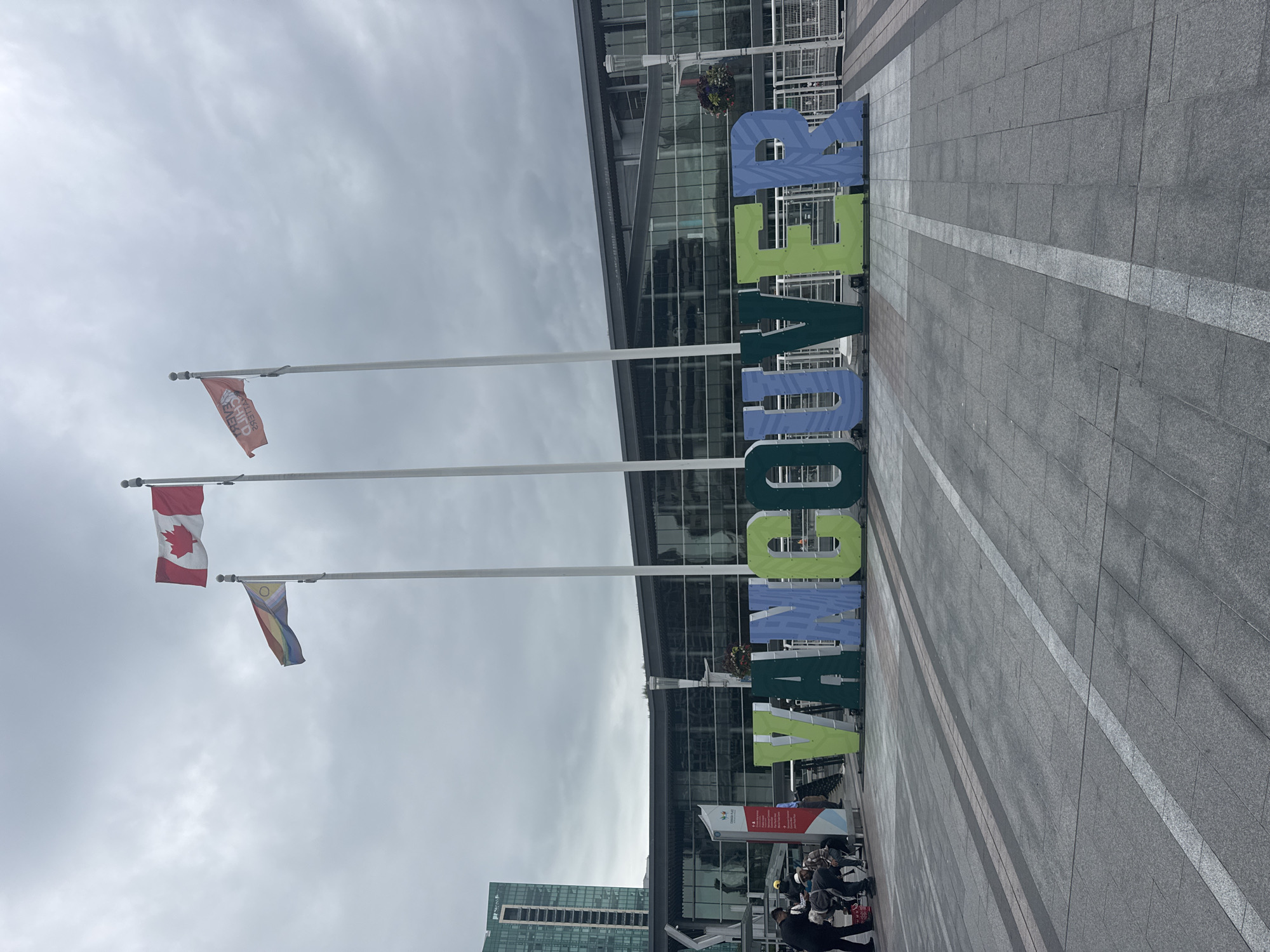 The image shows a large, colorful "VANCOUVER" sign in the foreground. The letters are green, blue, and light green, with a striped pattern on the blue letters. Behind the sign, a modern building with large glass windows is visible. Three flags are flying on poles to the left of the sign: the Canadian flag, a flag with rainbow stripes and an Indigenous symbol, and a flag with red text. Several people are gathered to the left of the sign, some appearing to be looking at a pamphlet or map. The ground in front of the sign is paved with large, grey square tiles. The sky is overcast with gray clouds. The scene appears to be an outdoor public plaza in Vancouver, Canada, likely in front of a convention center or a large public building.