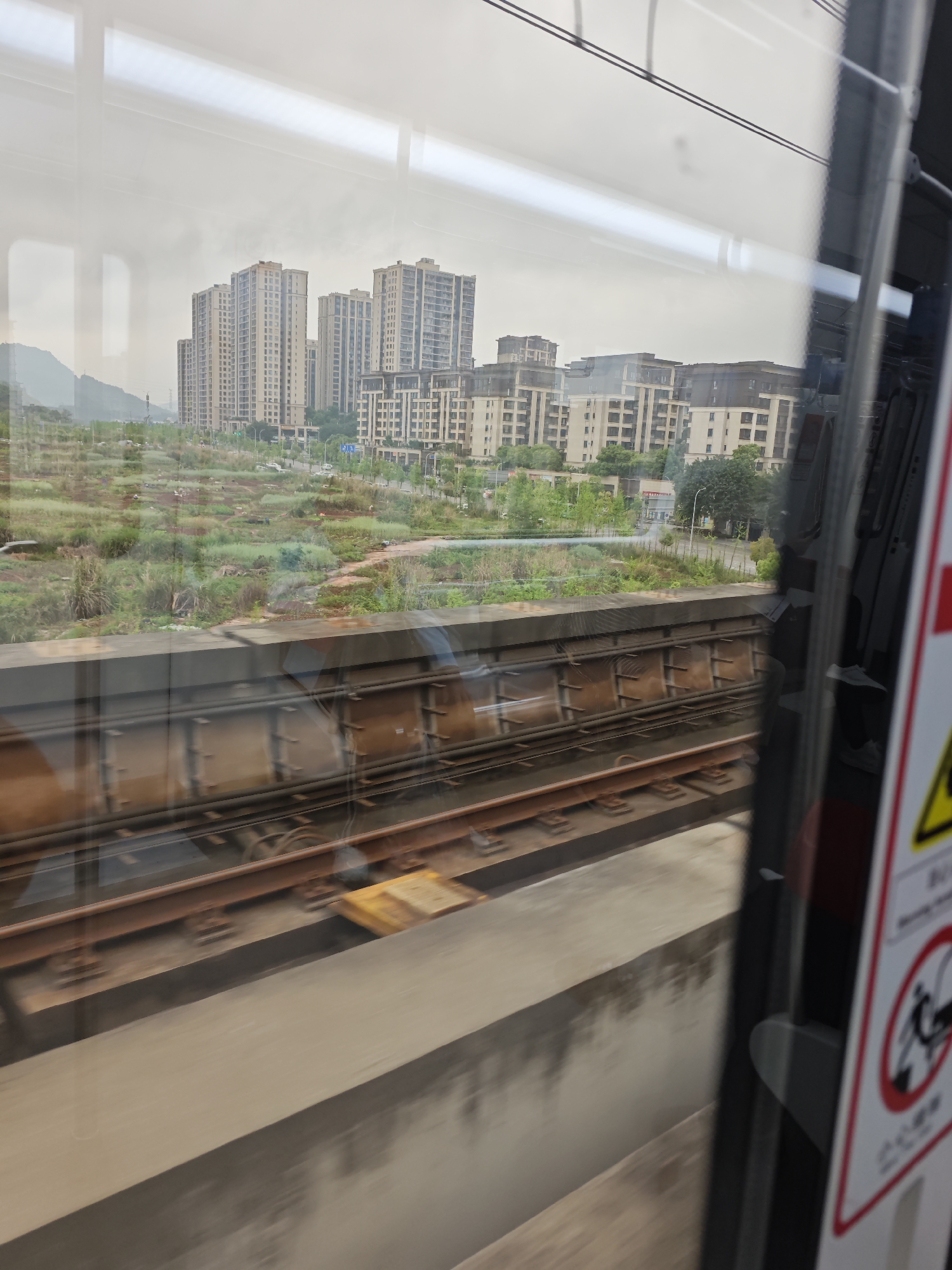 The image shows a view from a moving train, likely in the Chongqing High-tech Zone, China. In the background, there are several residential buildings, ranging from high-rise apartments to lower, multi-story complexes. In front of the buildings, there is a field with some vegetation. The foreground features the train tracks and the interior of the train, including a warning sign on a door. The weather appears overcast or foggy.