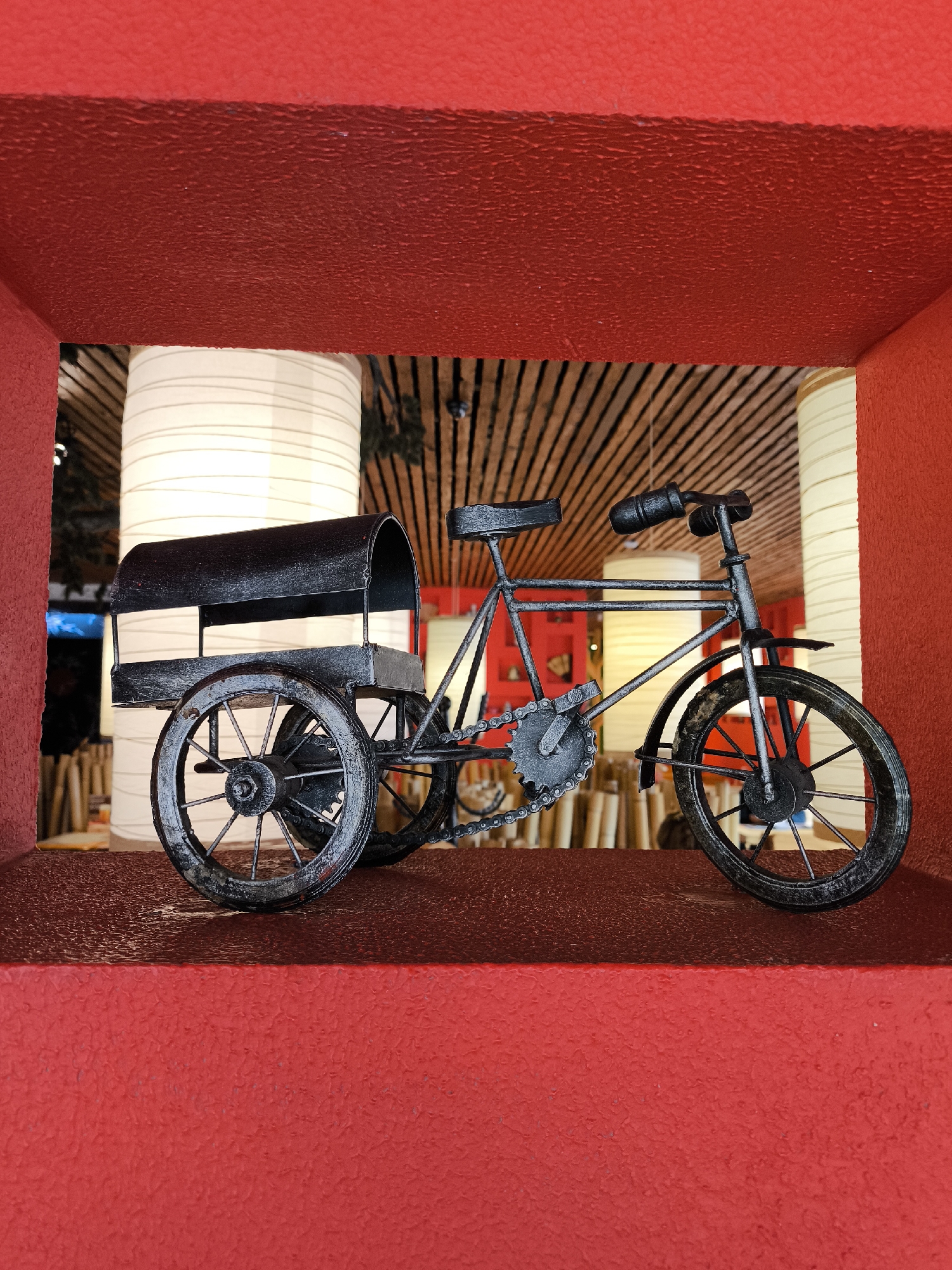 A metal tricycle figurine is positioned within a red cubby. The background contains paper lanterns hanging from a wooden slatted ceiling. The tricycle is facing right.