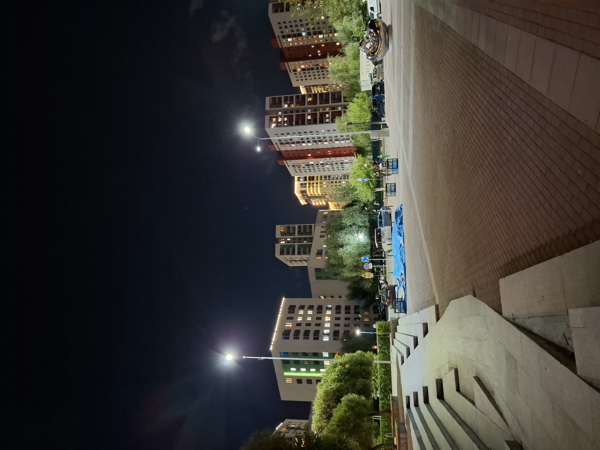 This image captures a nighttime scene in Tyumen, Russia, likely a residential area or public square. A modern apartment complex with lit windows dominates the background. In the foreground, a paved area with brickwork meets a concrete staircase and plaza. Several cars are parked on the street, and a few people are visible near some street furniture. A large, abstract metal sculpture is also present. The sky is dark, with a streetlamp illuminating the area.