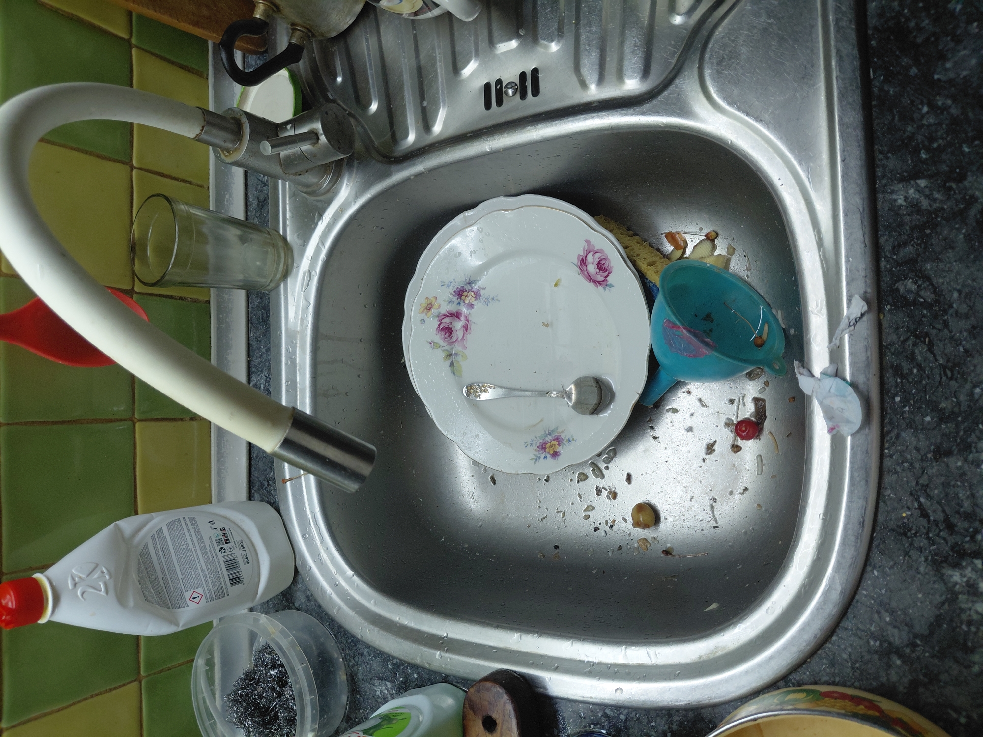The image depicts a dirty kitchen sink in a residence located in Marynivka, Ukraine. The sink is filled with dirty dishes, food scraps, and used kitchen utensils. A white bottle of dish soap and a container of steel wool are visible on the counter behind the sink. A glass of water sits on the counter next to the faucet. The sink area is cluttered and appears to be in a state of disarray.