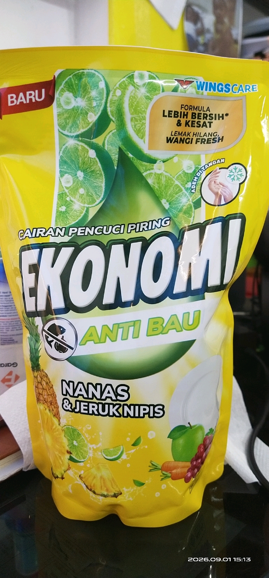 The image shows a close-up of a yellow pouch of dish soap. The brand name, "EKONOMI," is prominently displayed in large white letters with a black outline. Above the brand name, it says "CAIRAN PENCUCI PIRING," which translates to "dishwashing liquid." The pouch features images of limes, a pineapple, and what appear to be lemons. There is also text in an orange oval that says "FORMULA LEBIH BERSIH* & KESAT" and "LEMAK HILANG, WANGI FRESH." On the right side of the pouch, there is an image of hands with a snowflake symbol and text that says "ADEM DI TANGAN." Below the "EKONOMI" logo, it says "ANTI BAU," meaning "anti-odor." The bottom portion of the pouch displays "NANAS & JERUK NIPIS," indicating pineapple and lime as the scent. Scattered around the pouch are small green leaves and water splashes. A timestamp at the bottom right corner reads "2026.09.01 15:13." The pouch is placed on a reflective surface, possibly a table, and the background is out of focus, showing a blurred yellow object. There are no people visible in the image. The image is not taken in Tamiang Layang, Indonesia; it is a product photo.