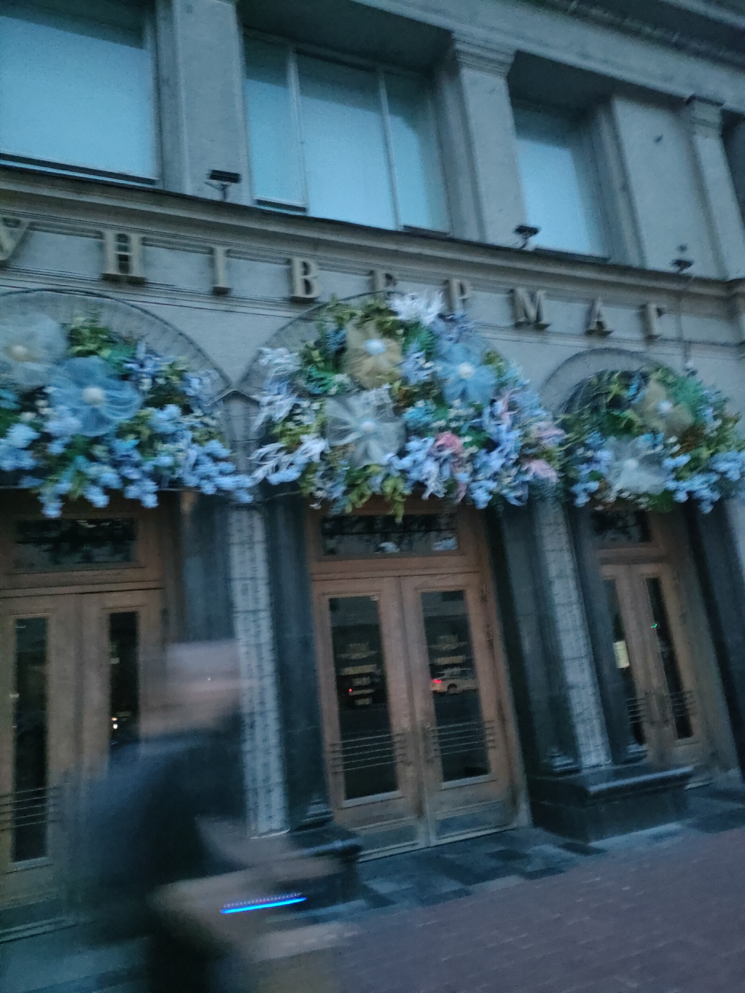 The image shows the facade of a building with large windows and ornate architectural details. Above the entrance doors, elaborate floral decorations are displayed. The building's name is visible above the floral arrangements, with letters spelling out "УНИВЕРМАГ". This is a common term for a department store in Belarusian and Russian. The image appears to have been taken at dusk or in low light conditions, as indicated by the ambient lighting and the blur of a person in the foreground. The building's facade suggests it is a well-established establishment, likely a prominent shopping center or department store. The overall impression is one of an urban environment, possibly in a city with a certain architectural heritage.
The text "УНИВЕРМАГ" on the facade indicates that this is a department store. Given the architectural style and the term used, this is likely "Univermag Belarus" (Универмаг Беларусь), a well-known department store located in Minsk, Belarus. The building itself is a significant landmark in the city. The image captures the entrance area of this department store, decorated with festive floral arrangements, likely for a special occasion or season. The presence of a blurred figure in the foreground suggests that the image was taken while someone was moving, possibly passing by the entrance, or perhaps exiting the store. The lighting conditions suggest it is either early morning or late evening, and the overall atmosphere is somewhat subdued, reflecting the twilight hours. The cobblestone pavement in the foreground further adds to the urban character of the scene.