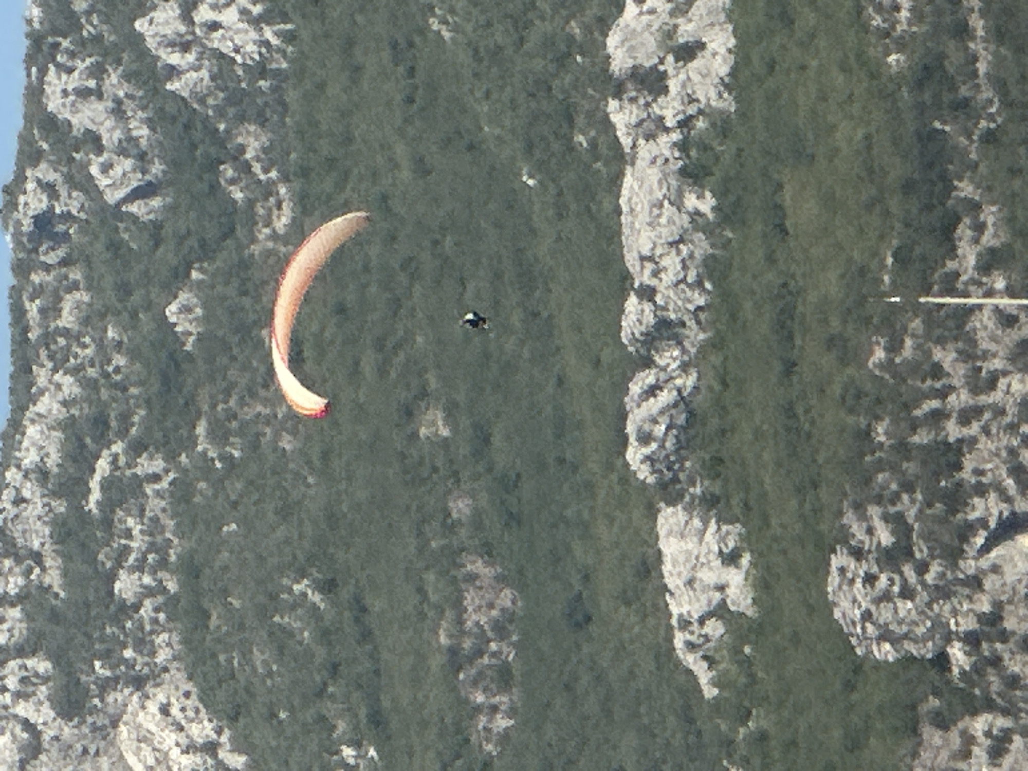 A paraglider is captured mid-flight against a backdrop of a densely vegetated mountain slope with rocky outcrops. The paraglider, with an orange and white wing, is positioned in the upper left quadrant of the image, with the pilot suspended below. The surrounding environment consists of a gradient of greens from the foliage and stark grey and white from the rocky terrain. The scene is likely part of the mountainous landscape near Boreti, Montenegro, a region known for its scenic vistas and opportunities for outdoor activities like paragliding.
