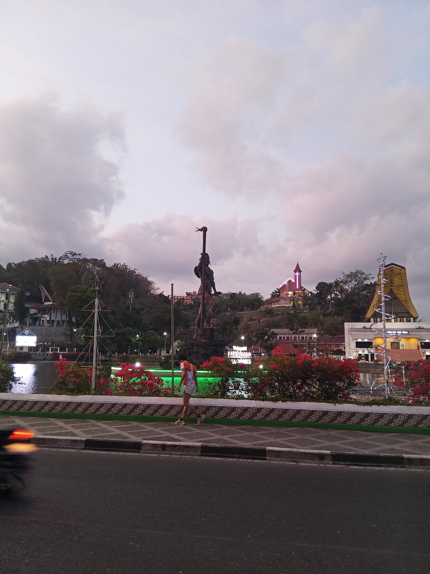 The image depicts a sunset scene in the city of Makale, Indonesia, focusing on the area around Lake Makale. In the foreground, a dark asphalt road is visible, with a sidewalk running parallel to it. The sidewalk is paved with patterned tiles and bordered by a low wall adorned with a decorative motif and green accent. A person, appearing to be a young woman wearing a white top and shorts, walks along the sidewalk. To the left of the sidewalk, a blur of motion suggests a vehicle, likely a motorcycle, passing by.
Behind the sidewalk, a body of water, Lake Makale, reflects the sky and surrounding lights. The lake is lined with vibrant red flowers and lush greenery. A prominent bronze statue of a warrior figure stands tall on a rocky base on the far side of the lake. Across the lake and on a hill in the background, several buildings are visible. These include a church with a prominent pink cross and a unique, traditional-looking building with a pointed roof, characteristic of Tana Toraja architecture. Christmas decorations, including a stylized Christmas tree outline illuminated with lights, are also present, suggesting a festive period. The sky is filled with dramatic, overcast clouds, with hints of pink and purple light indicating the approaching dusk. The overall scene portrays a lively urban waterfront at sunset, enriched by local landmarks and decorative elements.