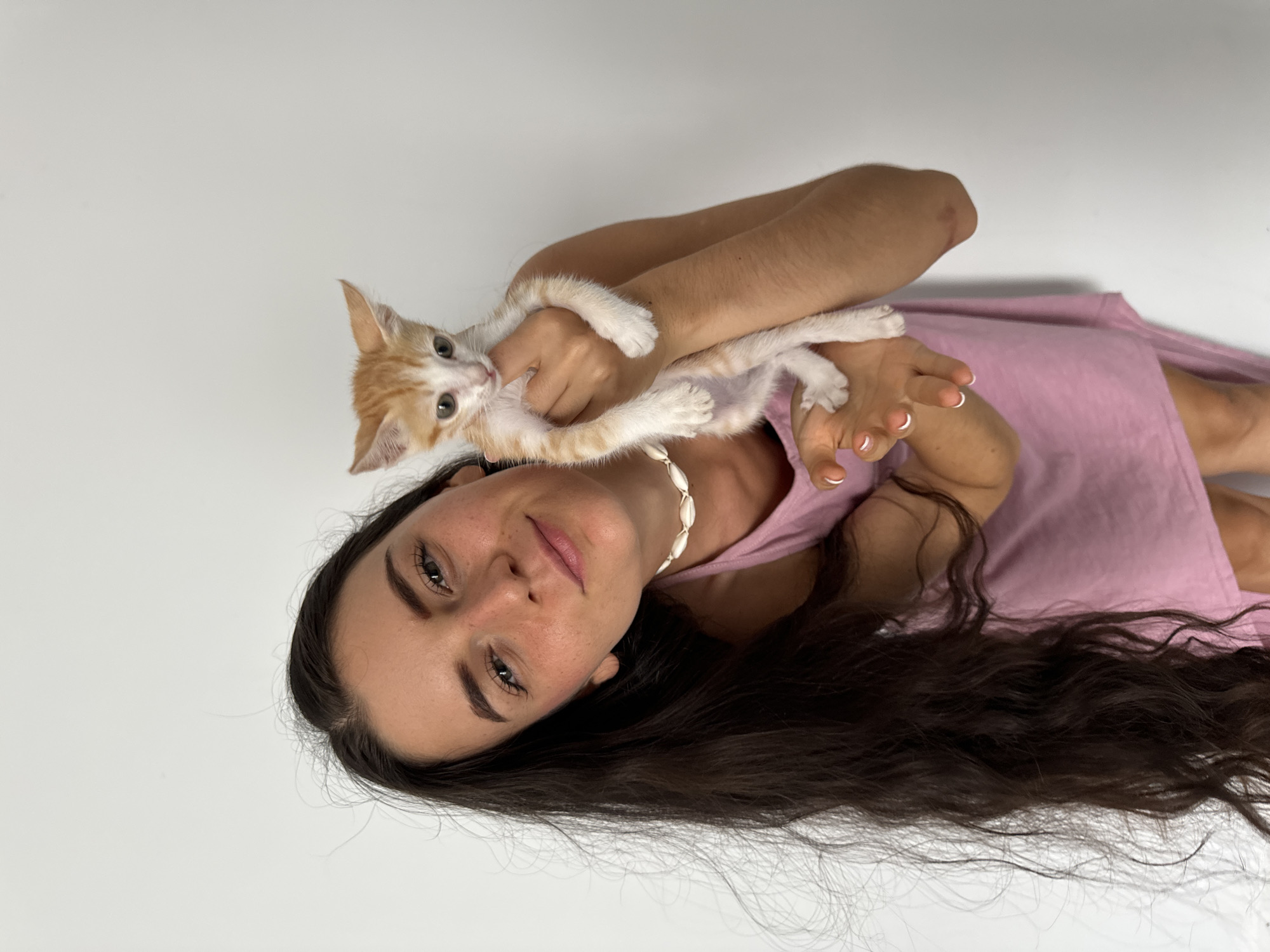 A young woman is depicted holding a small, orange and white tabby kitten. The woman has long, dark, wavy hair cascading to her left, light-colored eyes, and a natural complexion with visible freckles. She is wearing a light pink sleeveless top or dress and a white, shell-bead necklace. Her fingernails are painted with a French manicure style. She offers a gentle, warm smile towards the camera. The kitten, with light eyes, appears to be playfully engaging with her arms and is also looking towards the camera. The background is a plain, light-colored surface, suggesting an indoor or studio setting with bright, even lighting. The image is oriented diagonally, giving the impression that the subjects are looking up at the camera while lying down. Both the woman and the kitten convey calm and happy expressions. No text is visible in the image. The photo is set in the context of Mũi Né, Vietnam.