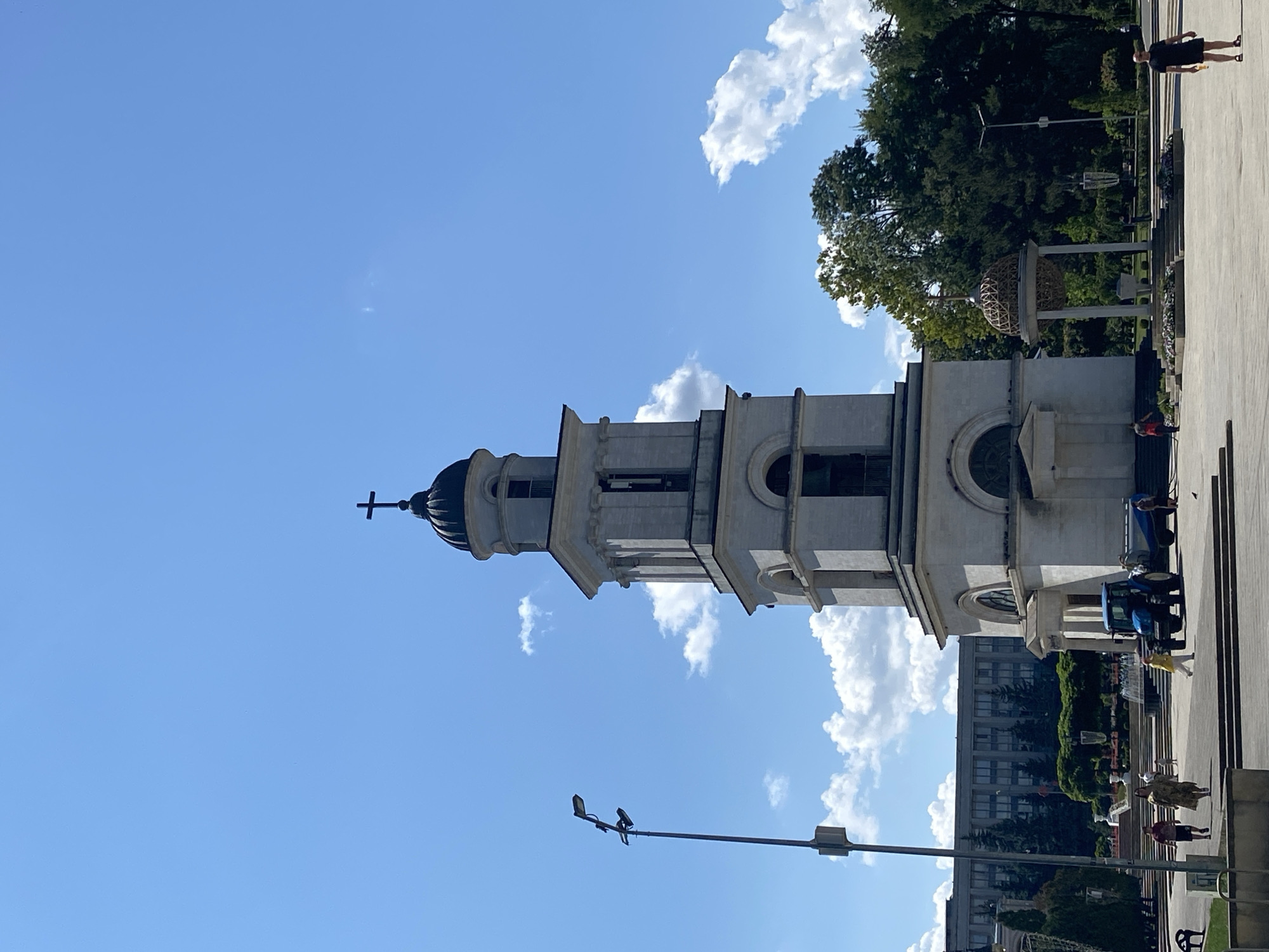 This is a photograph of the Bell Tower of the Nativity Cathedral in Chișinău, Moldova, taken on a sunny day. The sky is a clear blue with scattered white cumulus clouds. The Bell Tower, a tall, white stone structure with a dark dome topped with a cross, dominates the center of the frame. It features several tiered levels with arched openings and decorative cornices. In the foreground, a paved plaza is visible, with a few people walking. To the left of the tower, a tall lamppost stands. Behind the tower, a line of green trees and a large, modern building with multiple windows can be seen. To the right of the tower, a unique, spherical structure supported by columns is visible within a park-like setting. A blue tractor with a trailer is parked in front of the tower on the plaza.