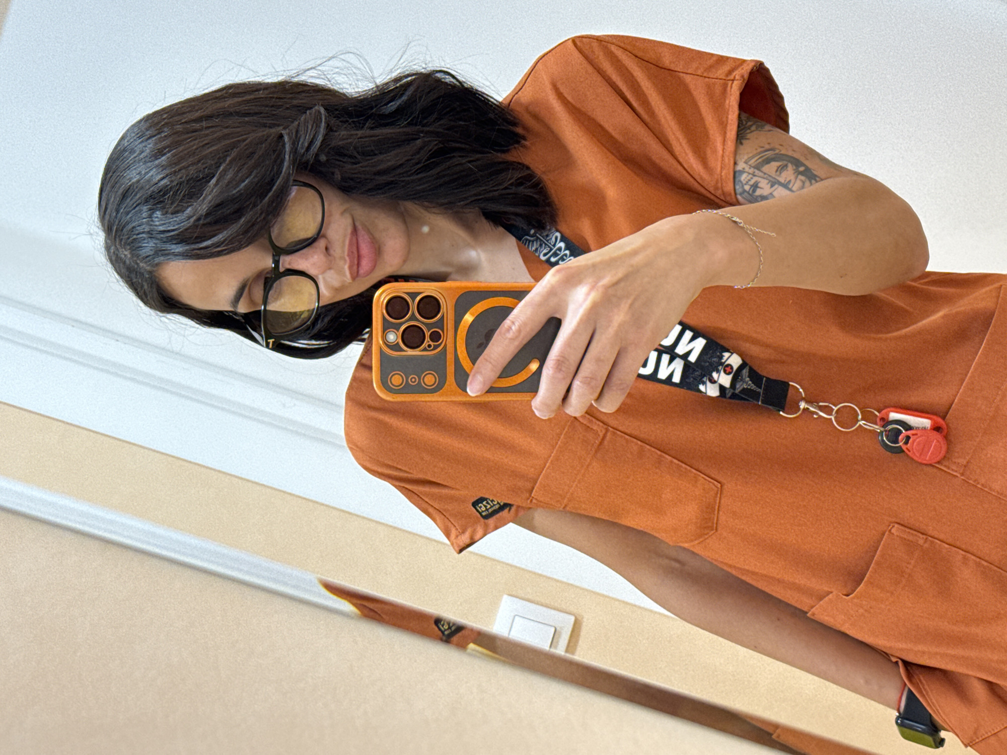 A woman is taking a selfie in a mirror. She has dark hair, is wearing glasses, and is dressed in orange scrubs. She is holding a mobile phone with an orange case. She is wearing a lanyard with a key attached. She has a tattoo on her left arm and a bracelet on her left wrist. The location appears to be indoors. No further information suggests it is in Pleven, Bulgaria.