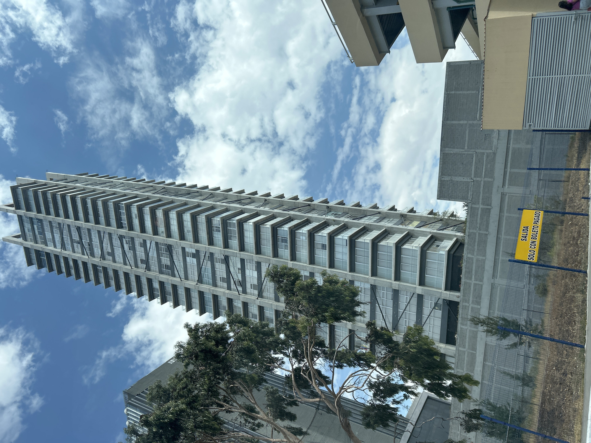The photograph shows a tall, modern building with numerous balconies against a partly cloudy blue sky. The building appears to be located in San Bernardino Tlaxcalancingo, Mexico, based on the sign written in Spanish which is visible on the right side of the image. Another building is partially visible behind trees at the base of the tall building. The photograph appears to have been taken looking up at the buildings.
