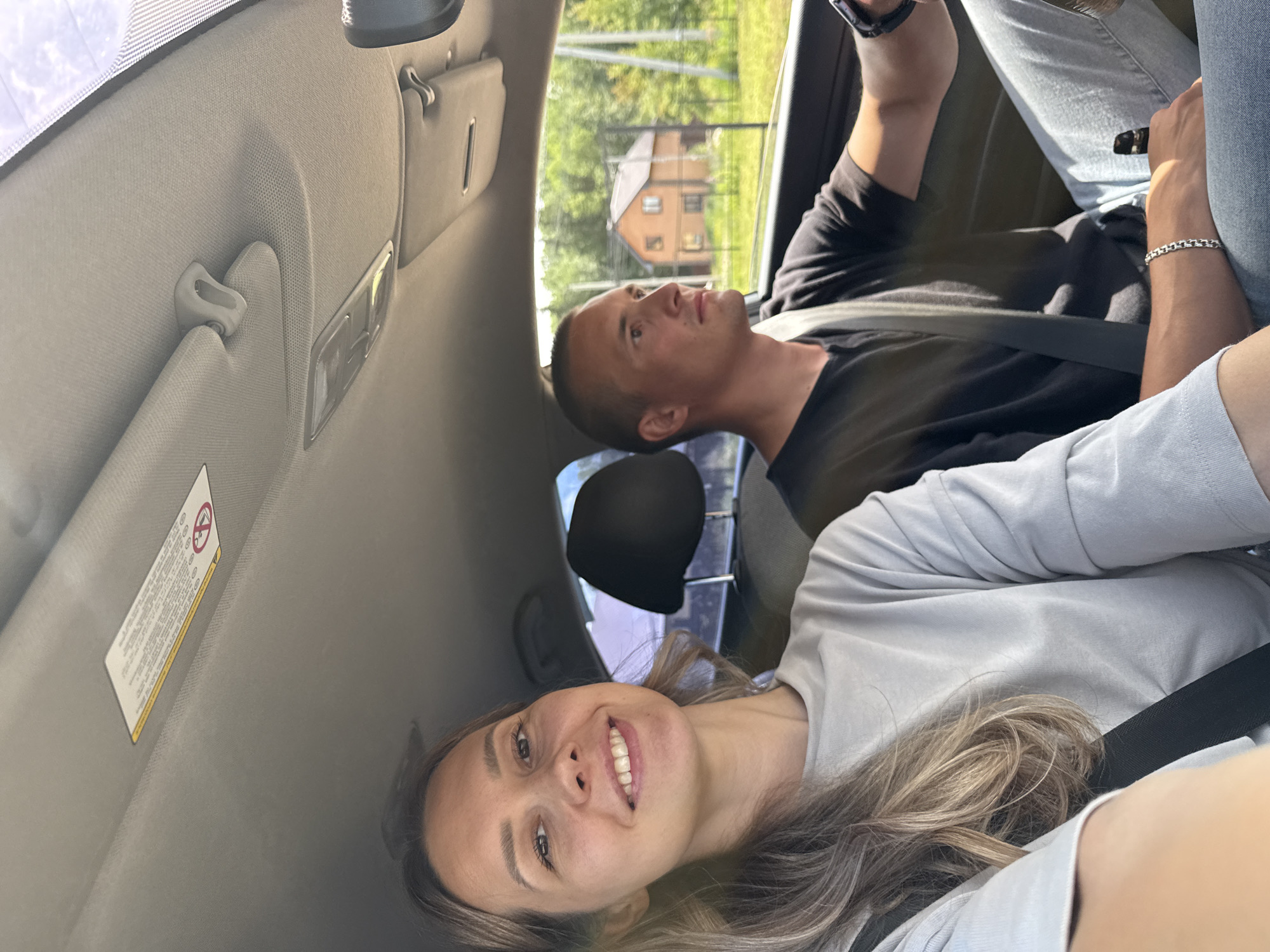 Two individuals, a woman and a man, are inside a car. The woman is in the foreground, smiling at the camera, with her hair falling over her left shoulder. She is wearing a light grey t-shirt and a black seatbelt. The man is in the passenger seat, looking forward and to the right, and appears to be driving. He is wearing a black t-shirt and a grey seatbelt. He has a silver bracelet on his left wrist. Outside the car, through the windows, a rural landscape is visible, with trees and a house with a red roof. This scene is likely taking place in Chaikovsky, Russia, given the visual cues of the environment and architecture.