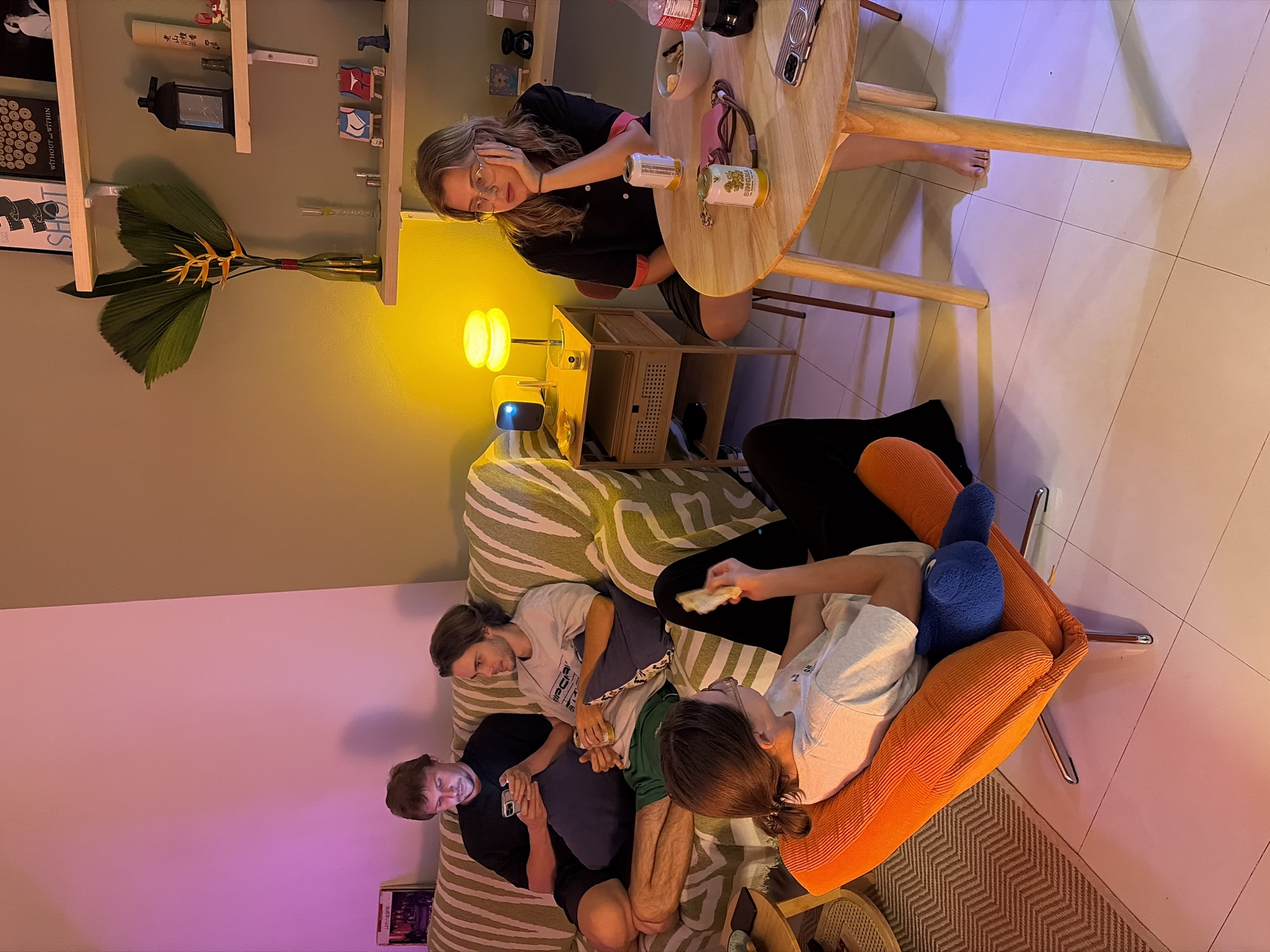 The image captures a casual evening gathering of five people indoors, taken from an elevated, rotated perspective. The setting is a cozy living space in Chang Phueak, Thailand, illuminated by warm yellow lamps and cooler ambient light, suggesting night or late evening.
In the upper portion of the frame, a woman with glasses and long hair sits at a small, round wooden table, resting her chin on her hand and looking towards the camera. On the table are two cans, one appearing to be a yellow Singha beer can, along with a bowl, a mobile phone, and a small pink bag. Beside the table, a bright yellow lamp on a wooden side cabinet provides warm light, near a small projector.
On the left, a multi-tiered wooden shelf is adorned with various decorative items, including a lantern, small figurines, a plant arrangement in a bottle, and books. Visible text on the shelf includes "WITHOUT WITHIN" and "SHOT," along with some illegible Asian characters.
In the lower part of the frame, four individuals relax on a sofa and an armchair. A person lies in an orange armchair, looking down, with a blue plush toy or pillow. On a striped sofa nearby, three men are seated. One with longish hair holds a can, another with shorter hair is engrossed in his phone, and a third, bearded man in a green t-shirt, rests comfortably. The floor is tiled, with a patterned rug partially visible. The overall mood is relaxed and social.
