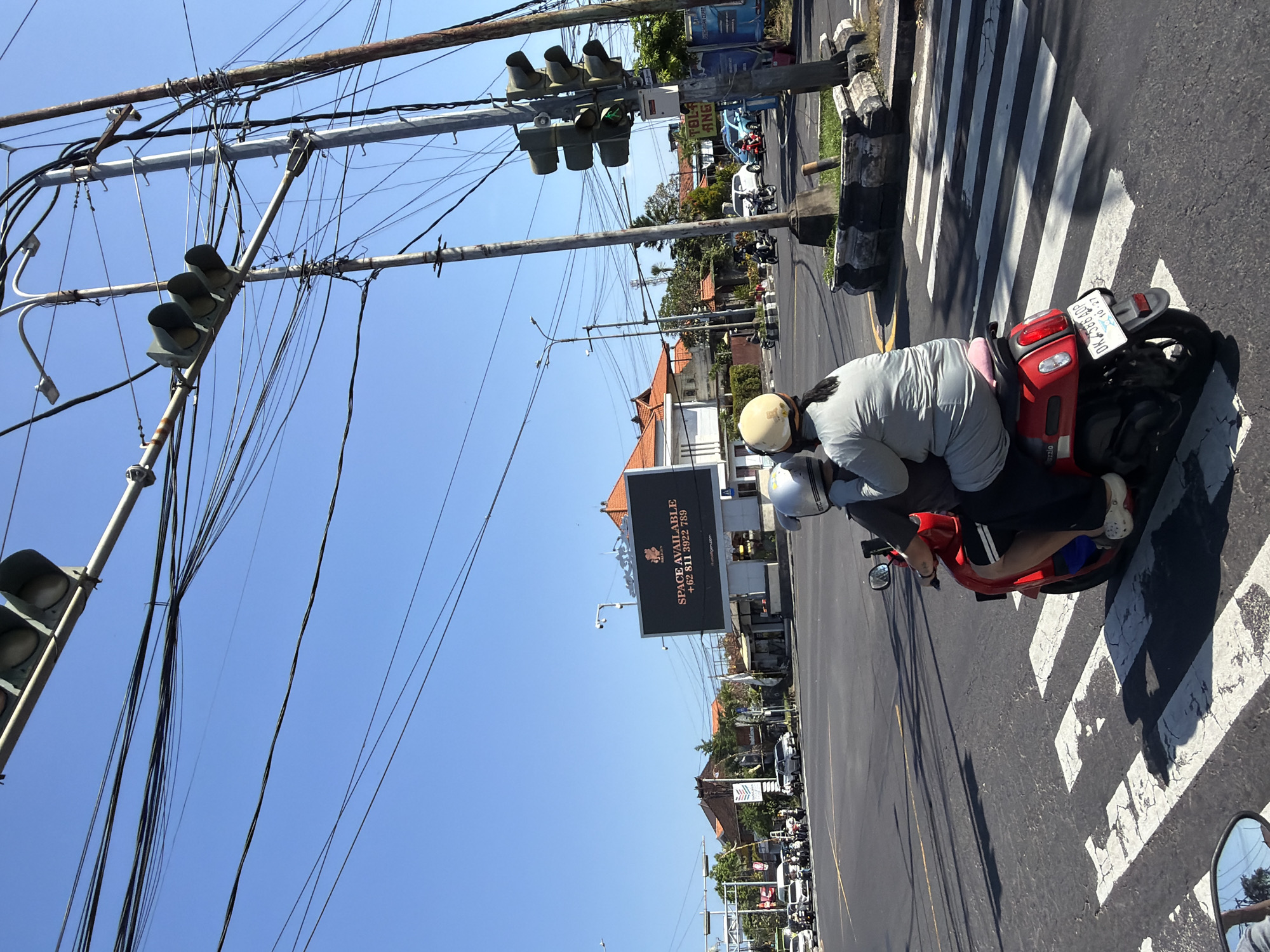 The image captures a street scene in Denpasar, Indonesia, with a bright blue sky overhead and a complex network of overhead electrical wires crisscrossing the frame. In the foreground, a red scooter carrying two people is stopped at a pedestrian crossing. The person in the back, wearing a grey top and dark pants, is leaning forward. The person in front, wearing a white helmet, is looking forward. Both are wearing helmets. The scooter has a license plate that reads "DK 3566 ADP".
Further in the background, traffic is visible at an intersection. Cars and motorcycles are present, and a traffic light hangs above the intersection. A large digital billboard on the right side of the road displays "SPACE AVAILABLE" and a phone number. Buildings with orange-tiled roofs are visible behind the billboard and along the street, characteristic of Indonesian architecture. The overall environment appears to be a busy urban intersection.