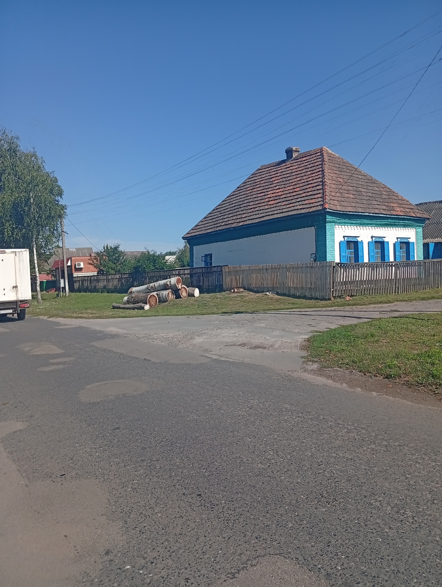The image depicts a scene in the village of Khomutets, Ukraine. A white truck is parked on the left side of the frame, facing right. In the background, a light blue and white house with a red tiled roof is visible, surrounded by a wooden fence. A pile of cut logs lies on the grass in front of the house. The sky is clear and blue, and the ground is covered with grass and a paved road. The overall impression is of a rural residential area on a sunny day.