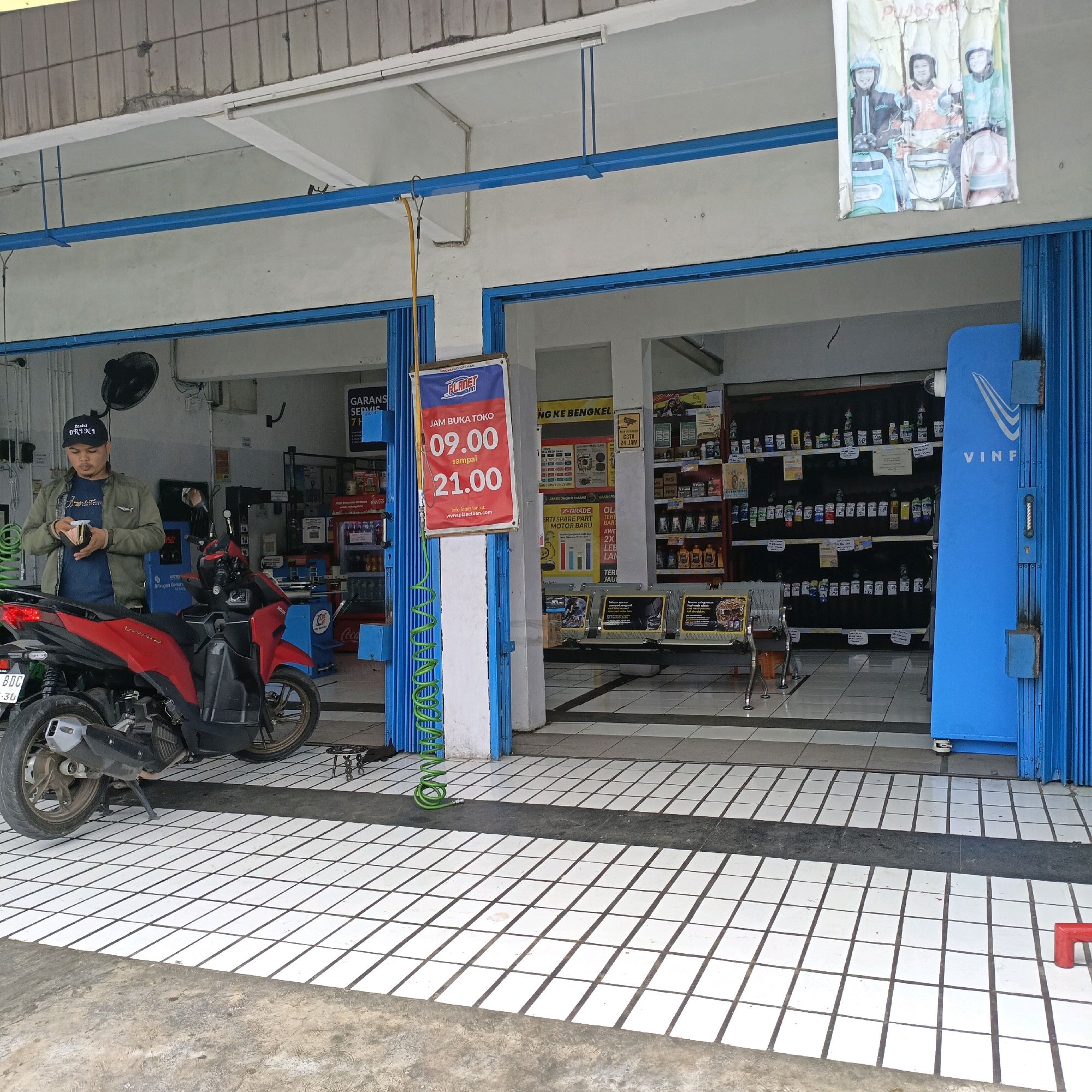 The image shows the exterior of a motorcycle repair shop in Salatiga, Indonesia. A red and black motorcycle is parked in front of the shop, with its rear wheel visible. A man wearing a dark t-shirt, a green jacket, and a black baseball cap stands next to the motorcycle, holding a small yellow object in his hands. The shop's entrance is open, revealing shelves stocked with various automotive products inside. A blue folding door is partially closed on the right side of the entrance, and a blue sign with white lettering that reads "VINF" is mounted on it. A poster with images of people in helmets is displayed above the entrance. The floor is tiled with white squares and black grout. A green coiled hose is visible on the ground near the entrance.