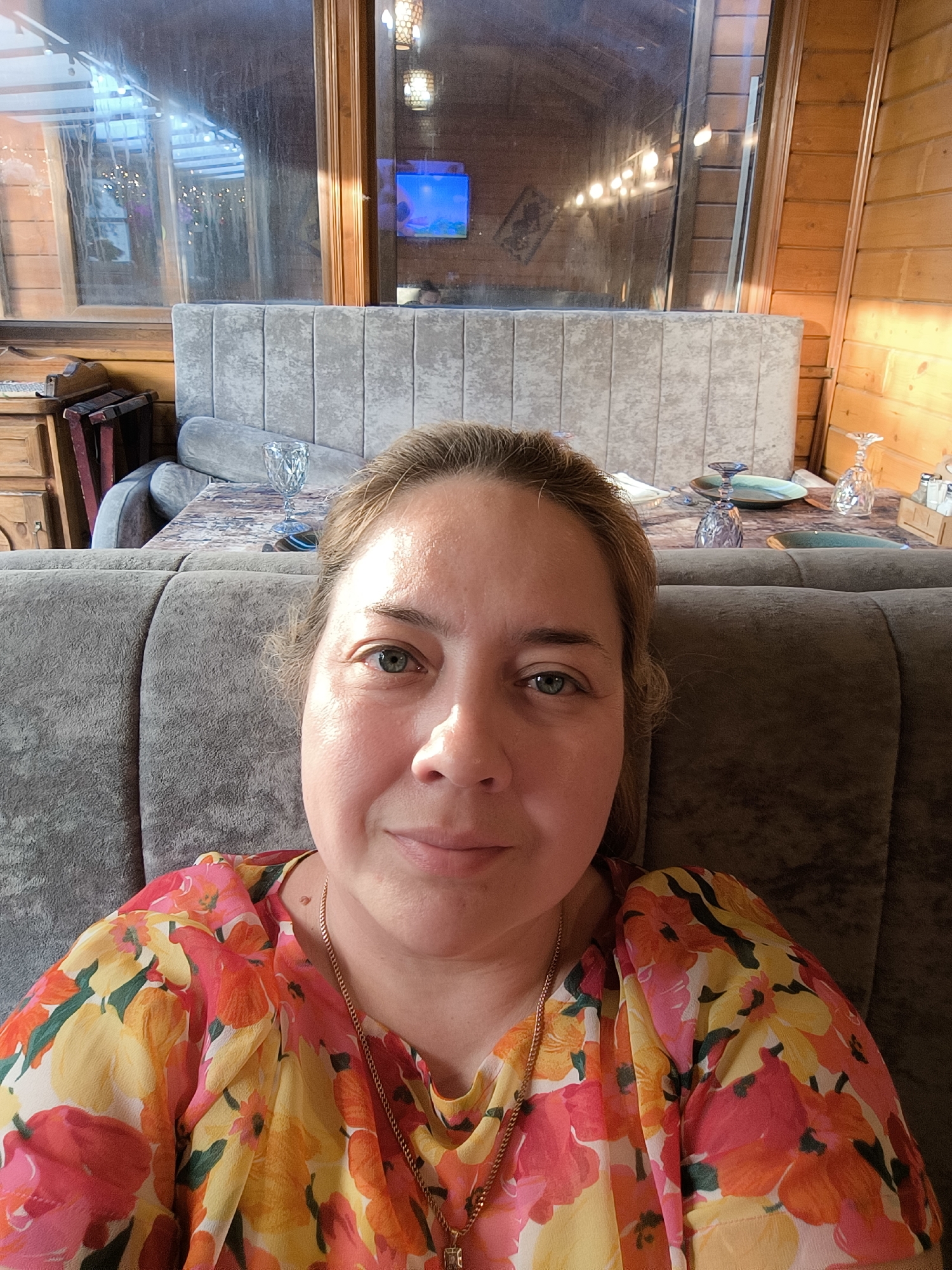 The image features a close-up selfie of an adult female in an indoor setting. The woman has light-colored hair, light-colored eyes, and a fair complexion, looking directly at the camera with a subtle smile. She is wearing a brightly colored floral top with patterns of red, orange, yellow, and pink, and a thin gold-colored chain necklace with a small pendant.
The background shows the interior of a dining establishment, characterized by light-toned wooden paneled walls. Large windows occupy the upper portion of the background, displaying reflections of outdoor elements including strung lights, structural components, and a blue-lit screen, possibly a television. A light gray, vertically ribbed upholstered bench or headboard-like structure is positioned behind the woman, separating her from a table.
On the table behind her, several items are visible: a patterned dark tabletop, an ornate clear glass goblet, a purple-stemmed glass, two ceramic plates (one turquoise, one off-white), a wooden serving board, and a wooden holder containing multiple small white condiment bottles or shakers. To the far left, a wooden side cabinet and parts of dark-colored wooden chairs are present. Lighting is a combination of ambient natural light filtered through the windows and warm artificial interior lighting, with a pronounced light source illuminating the wooden wall on the right side of the frame. The location is an interior space in Unknown, Unknown.