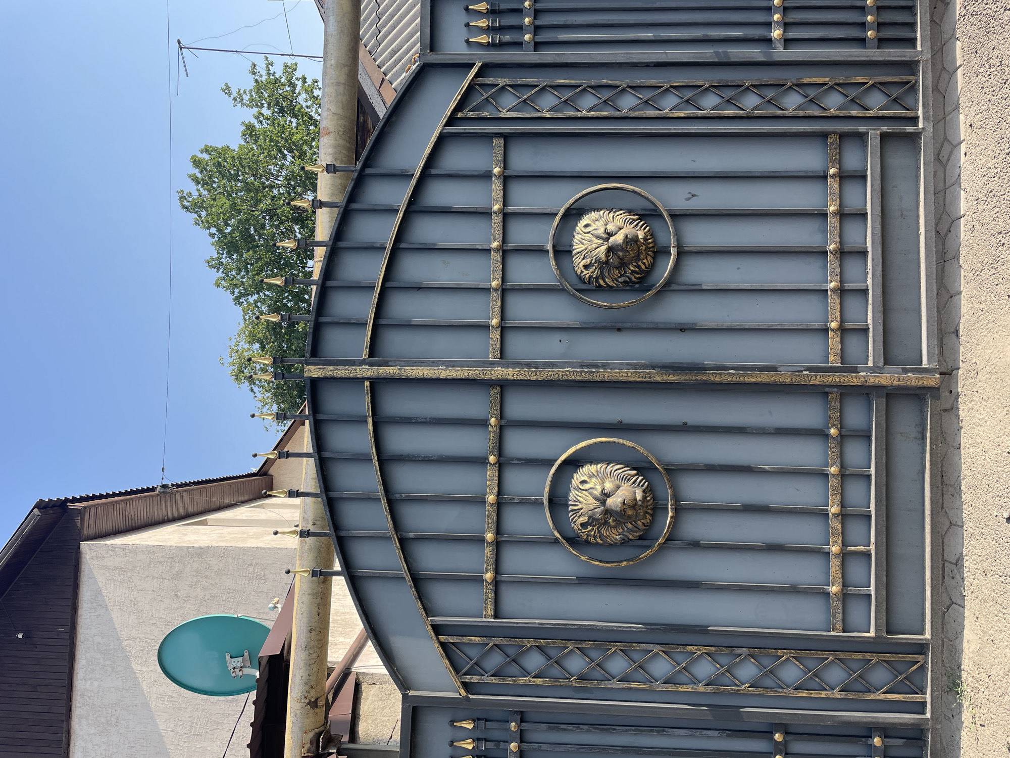 The image shows a close-up of a large, ornate metal gate in Almaty, Kazakhstan. The gate is a dark gray color with gold accents, featuring vertical bars and decorative scrollwork. Two prominent lion head medallions are mounted on the gate, one on each half, positioned at eye level. The gate appears to be part of a residential property, with a building and a satellite dish visible in the background. The sky is clear and blue, and a few trees are present. The ground in front of the gate is paved with interlocking stones.