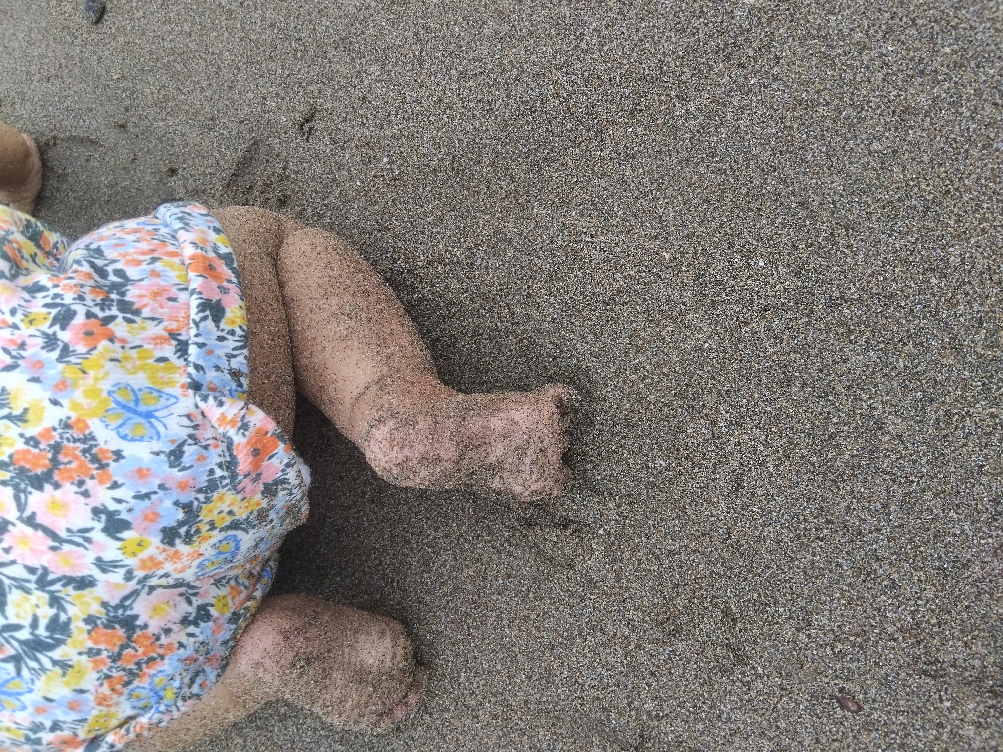 A close-up, ground-level shot shows a baby's legs and feet covered in wet sand on a beach in North Kuta, Indonesia. The baby is wearing a floral-print romper with white, yellow, orange, and blue flowers and butterflies. The baby's knees and feet are clearly visible and coated with sand. The background consists of the textured, wet sand of the beach.