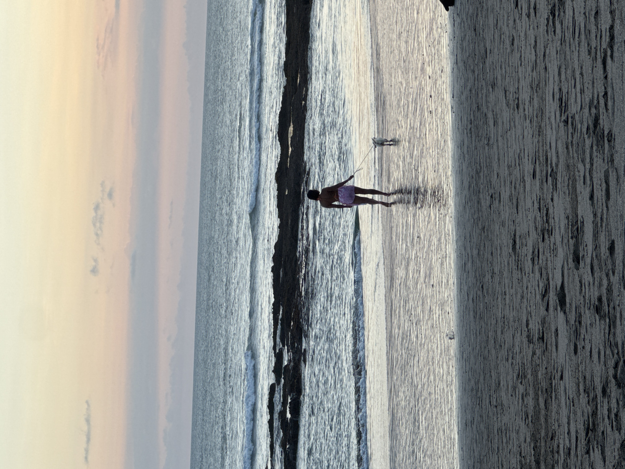 A woman walks a dog on the beach near a body of water. The location is the city of North Kuta, Indonesia.
