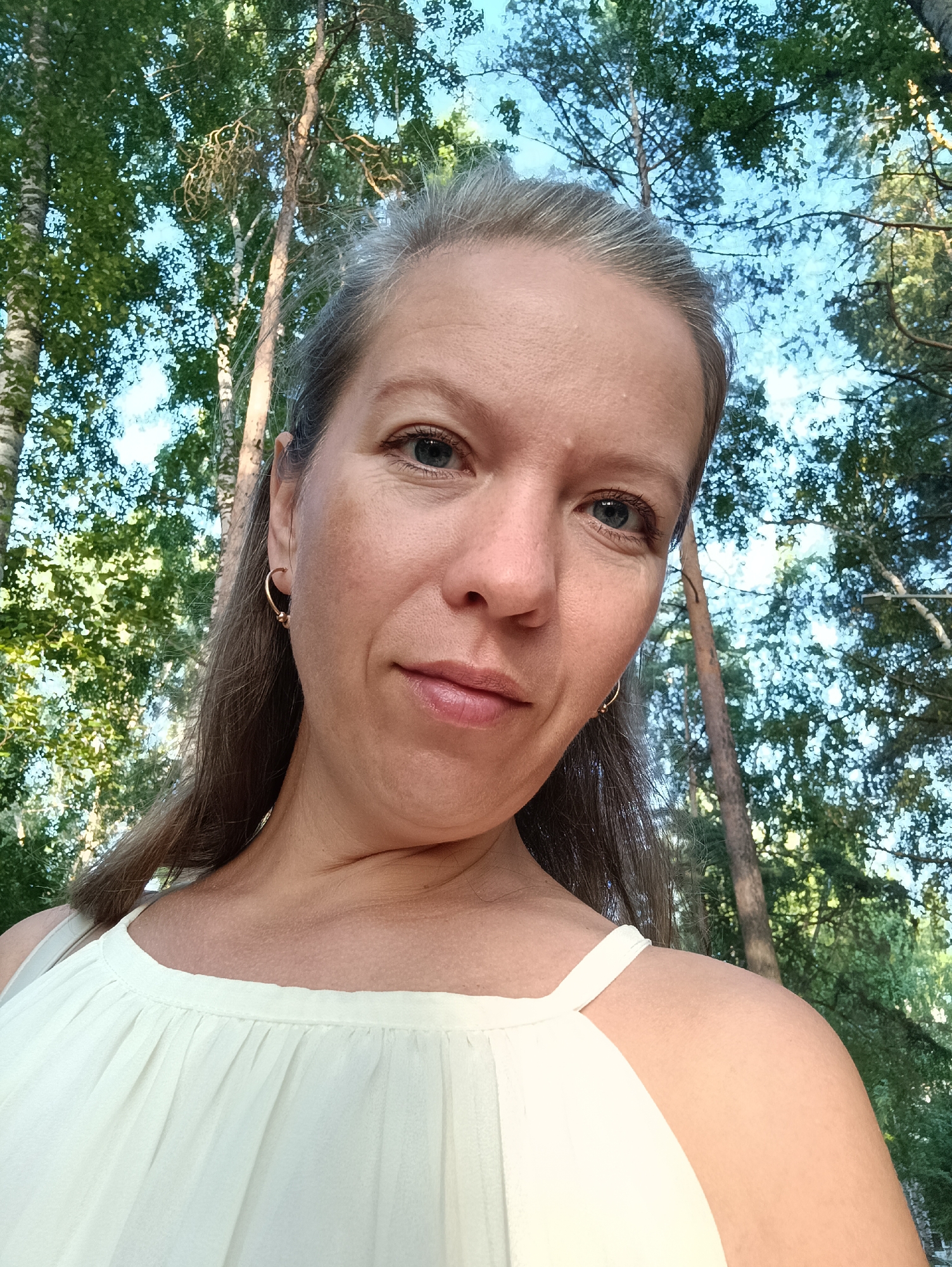 A woman with grey hair pulled back is looking at the camera, slightly tilting her head. She has blue eyes, light skin, and is wearing a light yellow top with thin straps. A gold hoop earring is visible on her left ear. The background is blurred with trees and a bright sky, suggesting an outdoor setting. The image appears to be a selfie taken in Chaikovsky, Russia.